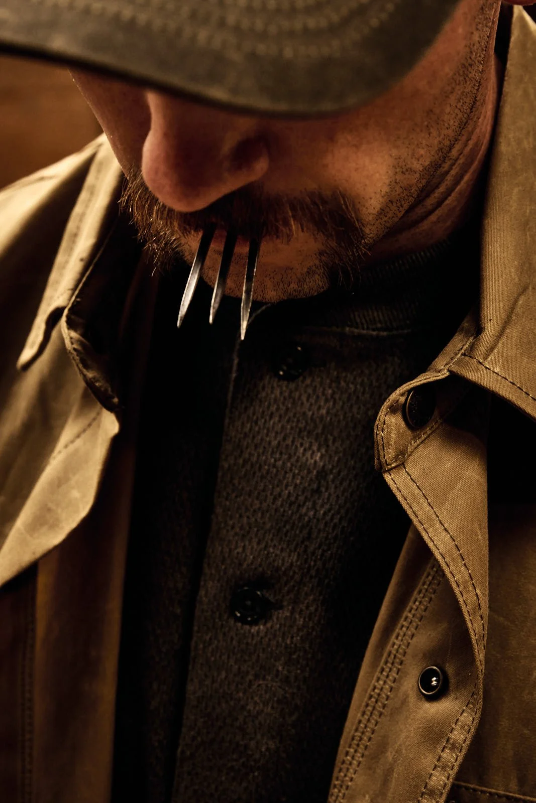 A man with a beard, and mustache, wearing a tan jacket over a dark textured shirt, with horseshoe nails in his mouth.