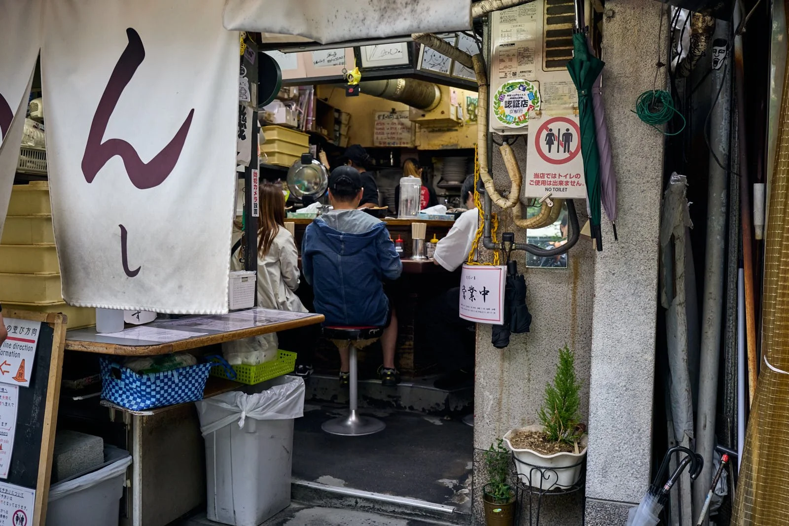 A small Japanese restaurant with customers seated at a counter, visible kitchen staff, and various signs and umbrellas around the entrance.