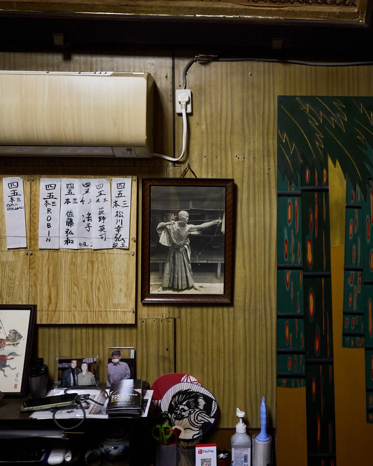 A wooden interior wall with photographs, Japanese calligraphy, and artwork. An air conditioning unit is mounted near the ceiling, with a power outlet and cord. There are various bottles, utensils, and photo frames on a desk or counter below, along wi