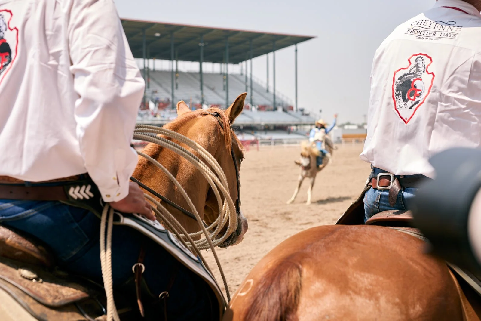 Riders on horseback at a rodeo event, with a focus on a rider wearing a white shirt with patches and another rider in the background riding a bucking horse.