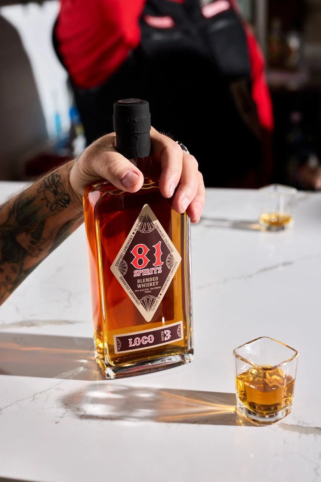 A person's hand is holding a bottle of 81 Spirits blended whiskey over a table, with a shot glass filled with whiskey in front of it and a blurred figure in a red and black jacket in the background.