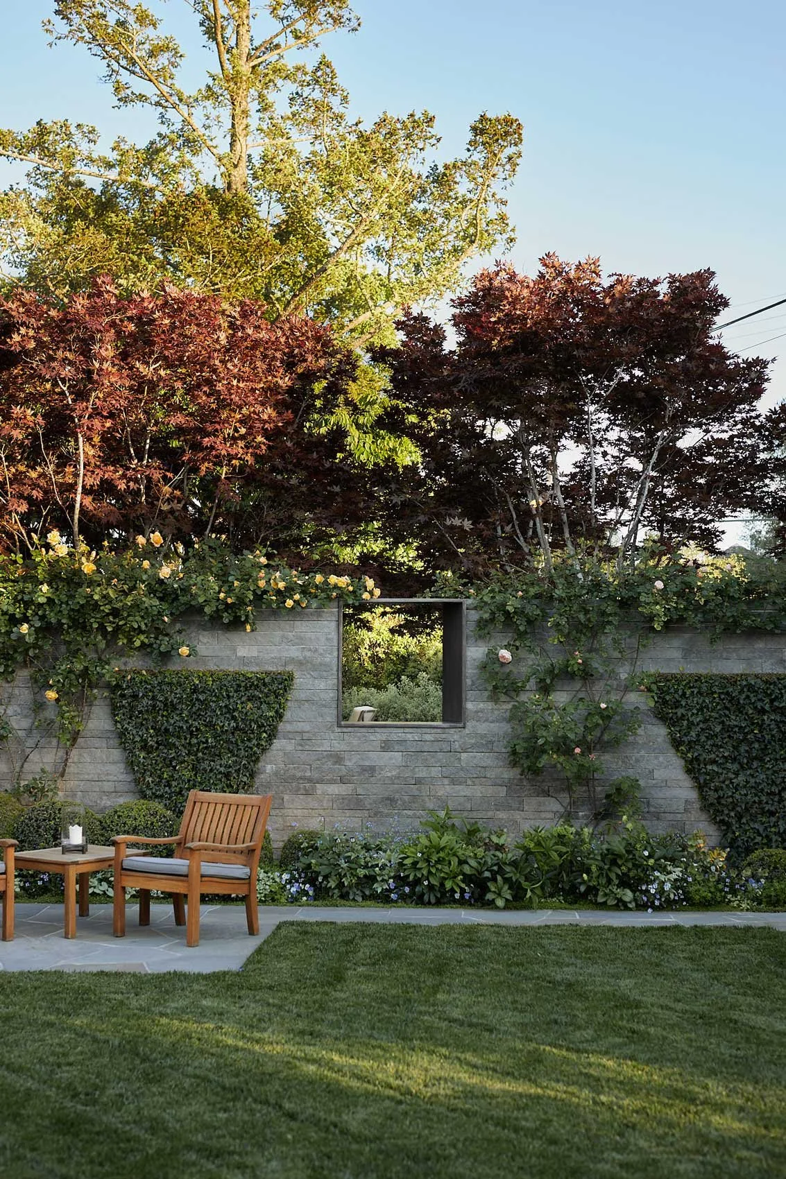 A backyard with a stone wall, various shrubs and trees, and a wooden chair and side table on a stone patio, with a grassy lawn in the foreground.