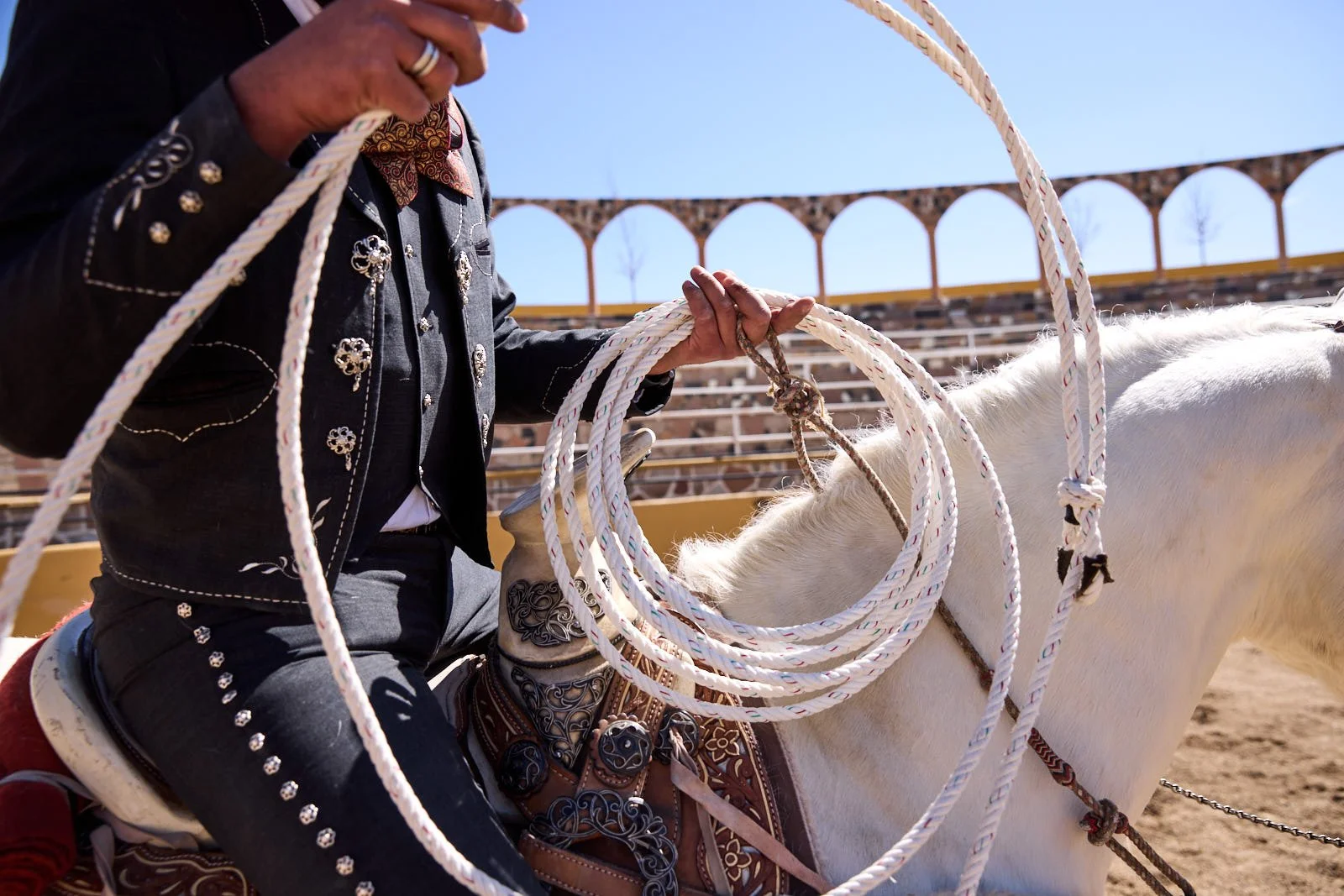 Close-up of a person in traditional Mexican attire, holding a lasso, riding a white horse in a rodeo arena with empty bleachers and clear blue sky in the background.