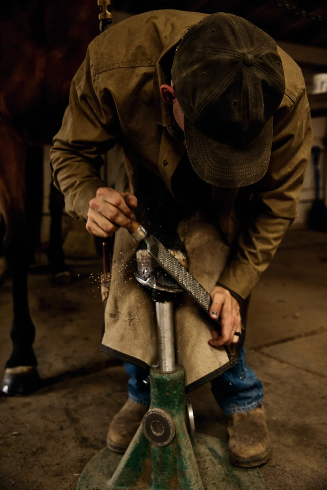 A person in work boots and a brown jacket, wearing a cap, is using a rasp file to smooth a horse hoof