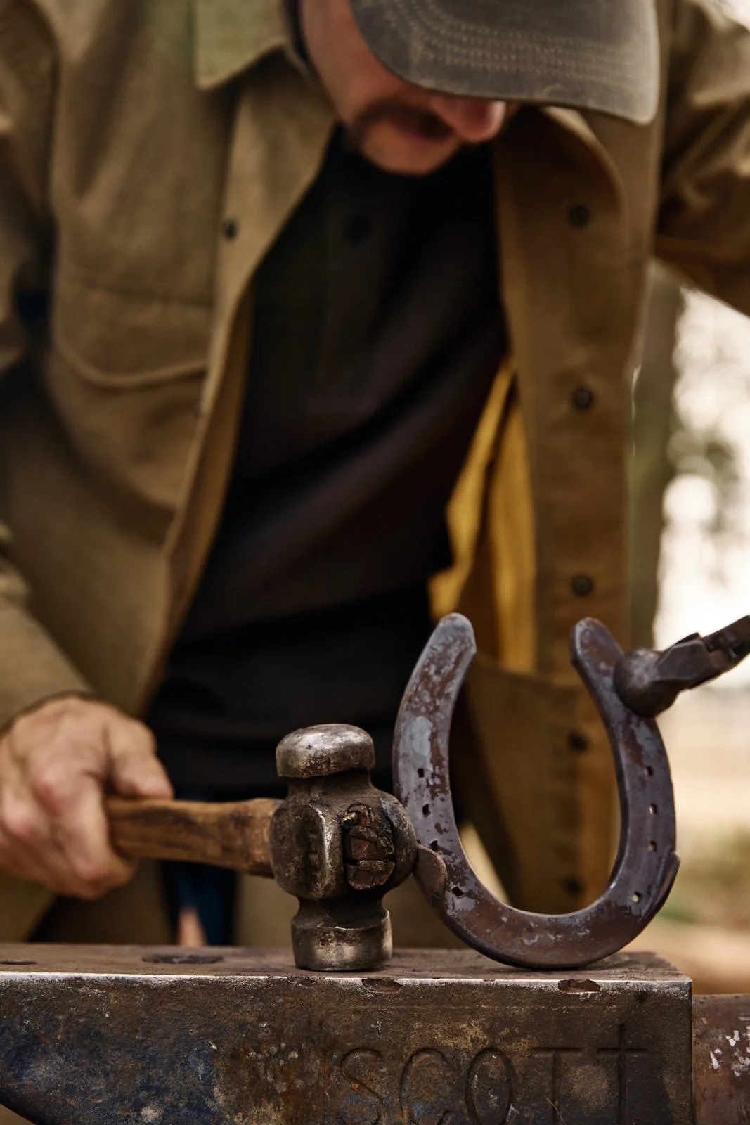 A man wearing a brown jacket and cap is forging a horseshoe with a hammer on an anvil.