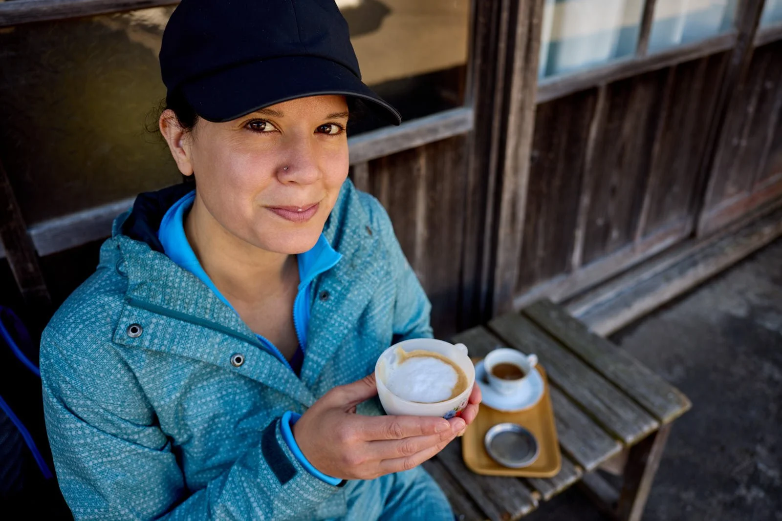 A woman in a blue jacket and black cap sitting outside, holding a cup of coffee with foam and caramel, with a small cup of coffee on a tray placed on a wooden bench.