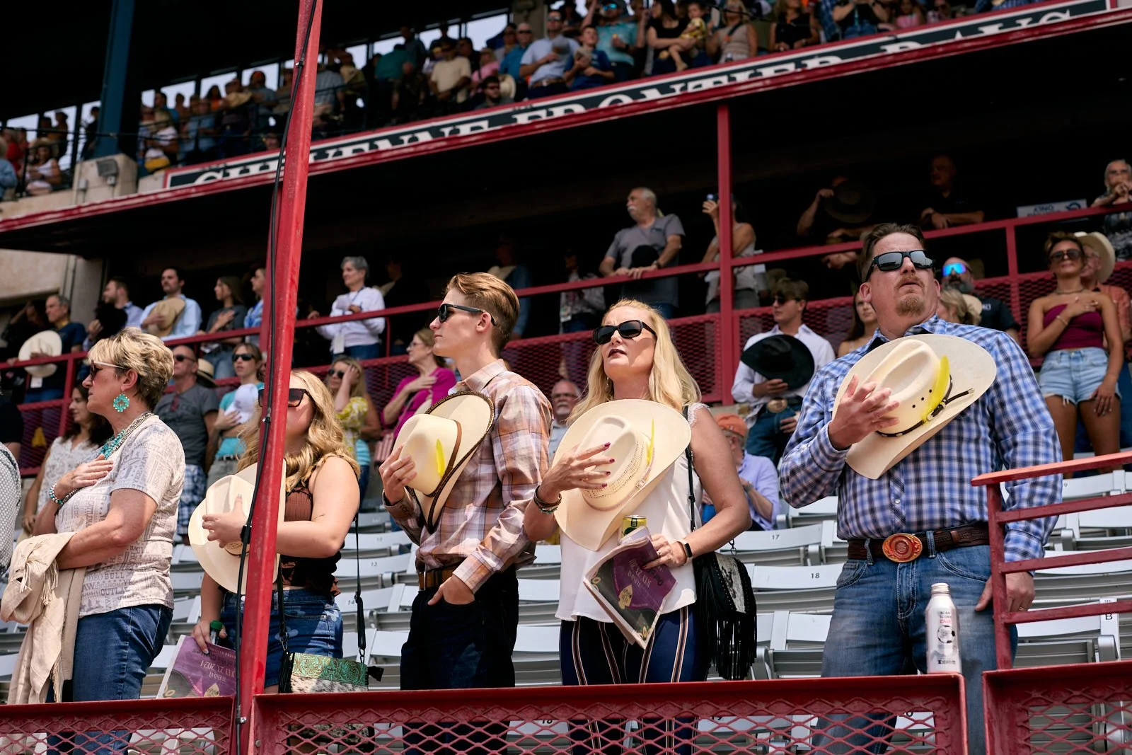 People standing in a row at a rodeo event, holding their hats over their hearts, with a crowd seated in the grandstand behind them.