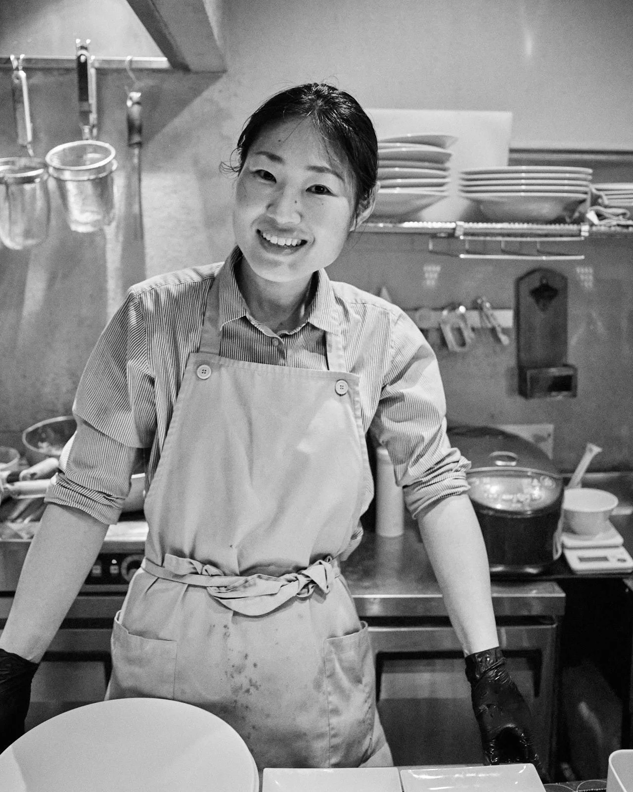 Black and white photo of a smiling woman in a kitchen wearing an apron, with dishes and kitchen utensils in the background.