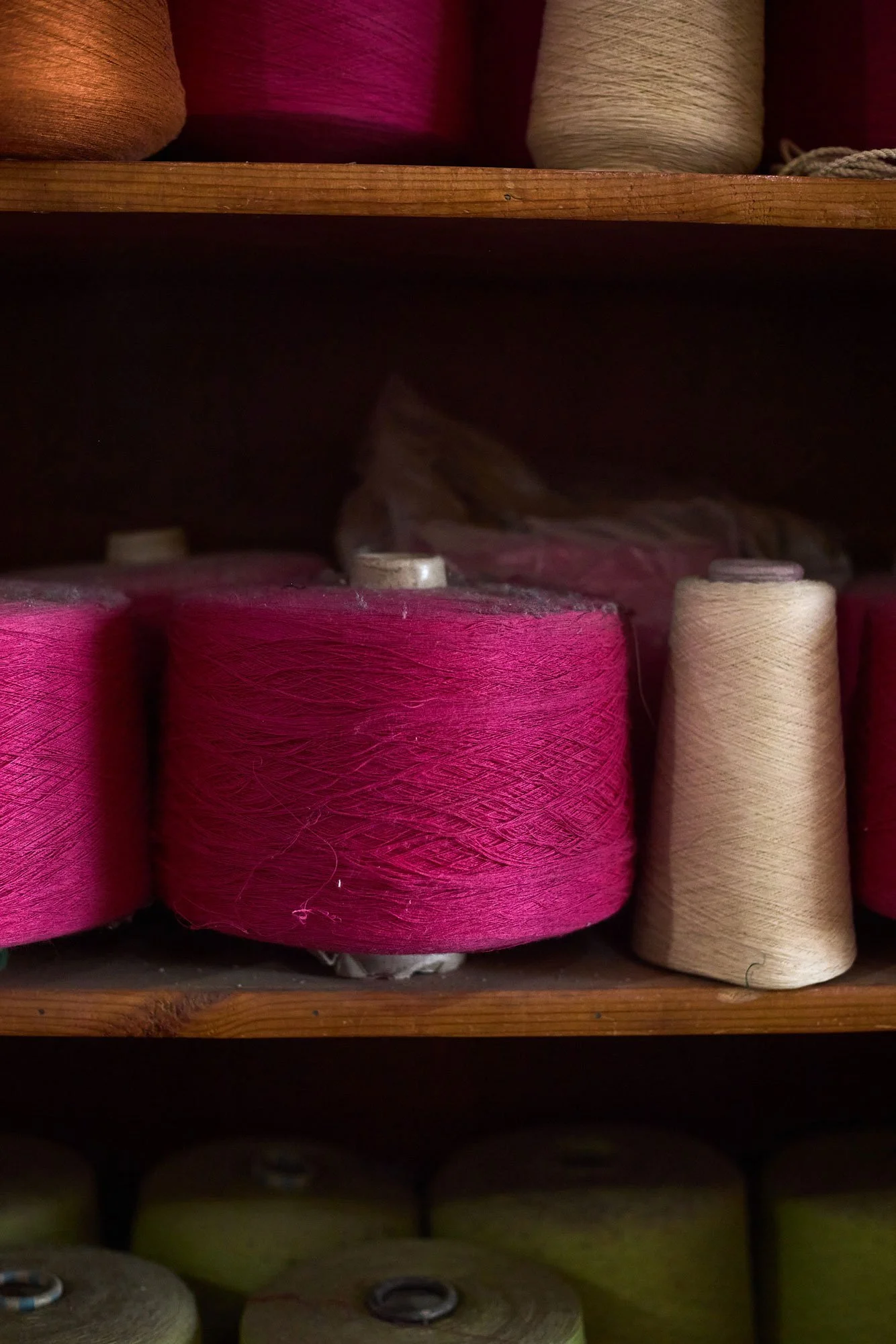 Spools of embroidery thread on wooden shelves, with pink and beige threads on the top shelf and various colors on the bottom shelf.