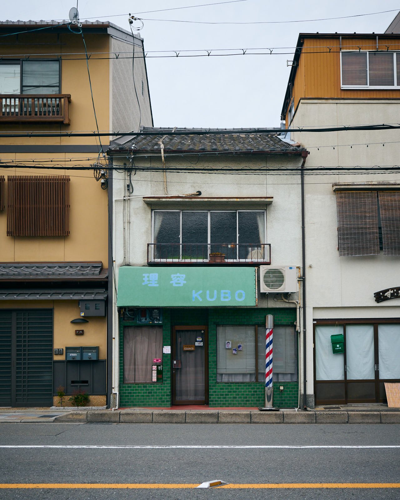 A street view of a multi-story building with various windows, balconies, and air conditioning units. The ground floor features a green storefront with a sign labeled "KUBO" and Japanese characters.