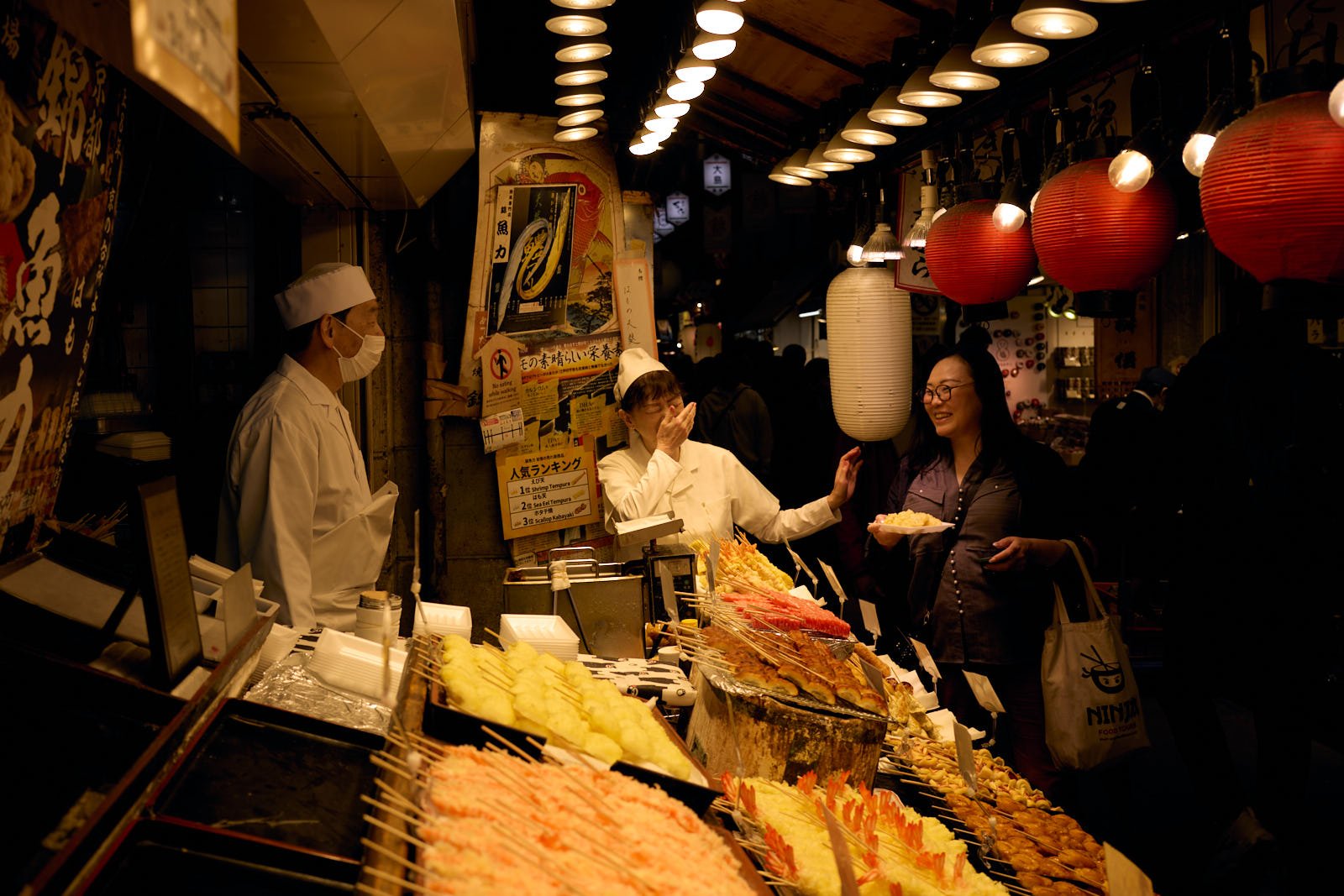 A lively night market scene with a food stall selling skewered tempura and other Japanese dishes. A smiling woman is buying food from a vendor, who is wearing a chef's outfit and face mask. Hot lanterns hang overhead, illuminating the busy atmosphere