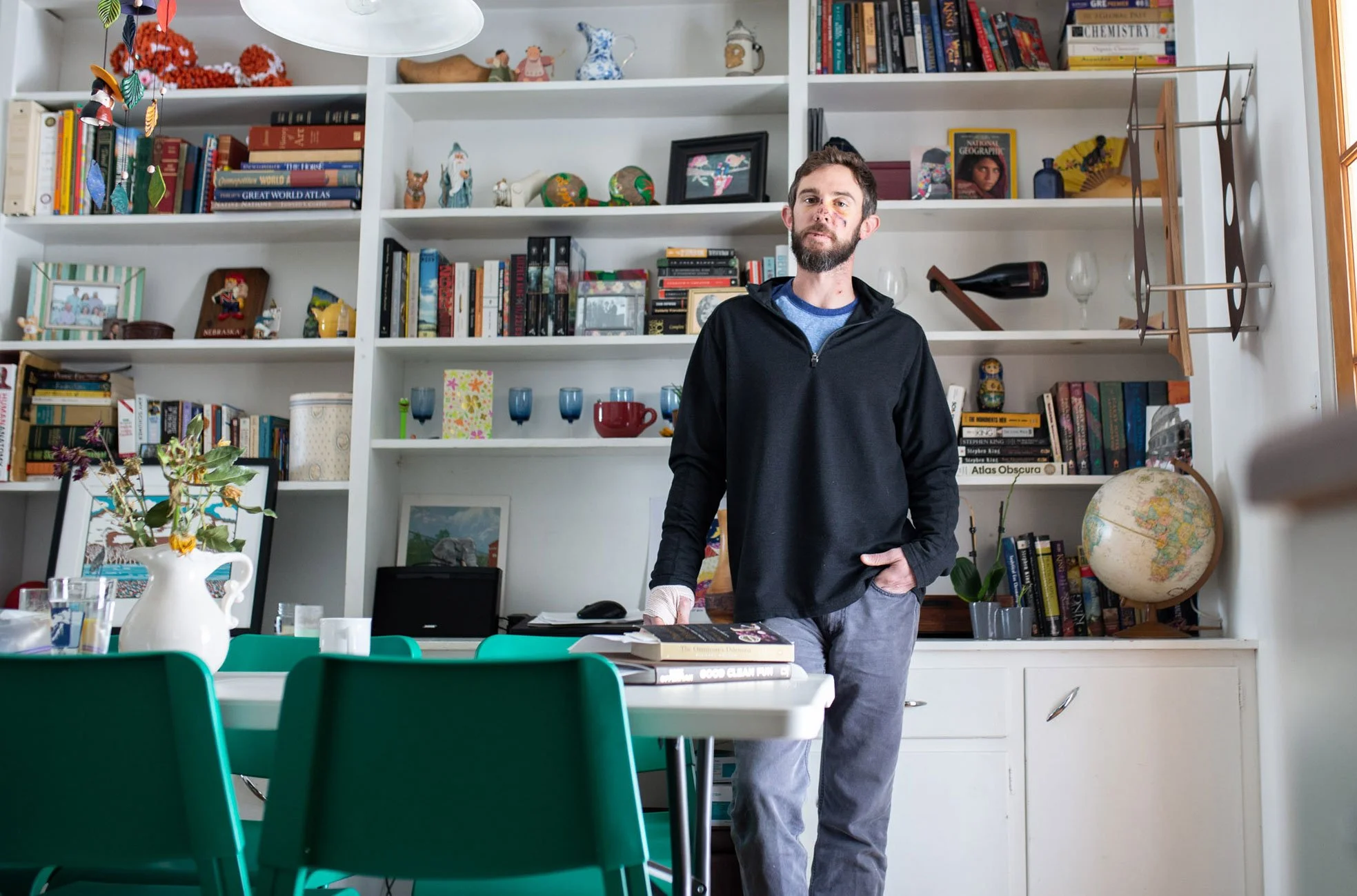 A man with a beard and casual clothing standing in a dining room with white shelves filled with books, decorative objects, and a globe behind him. The room has a table with green chairs and a floral vase.