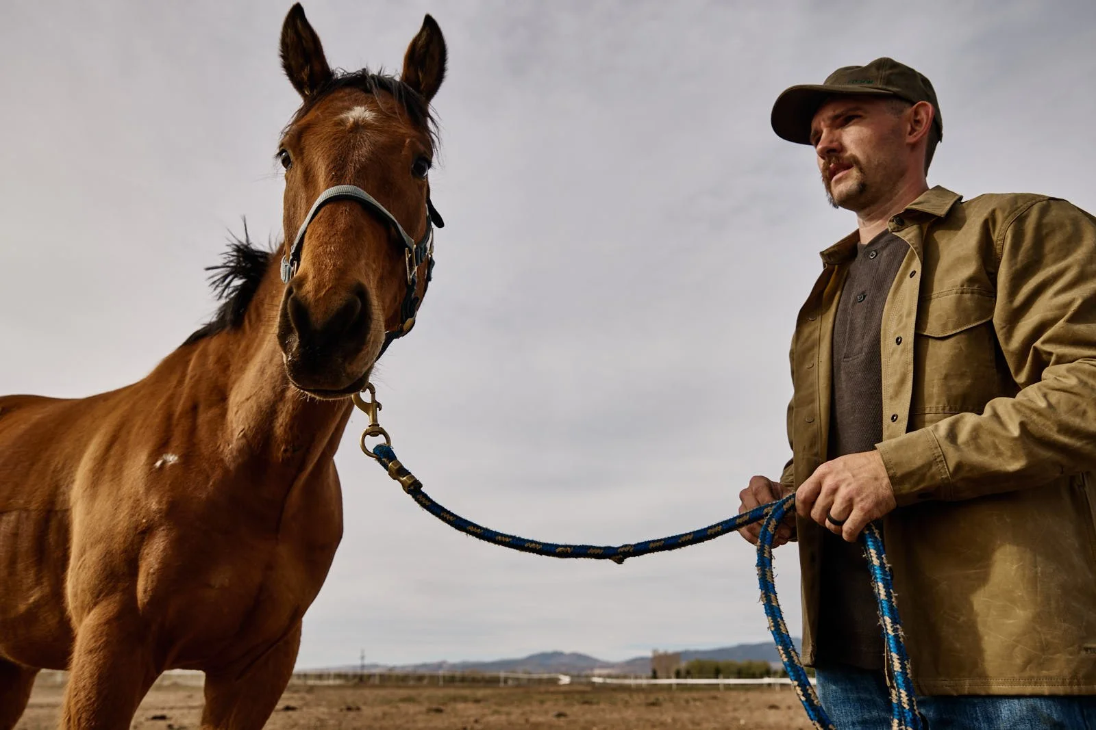 A man in a brown jacket and cap holding a blue lead rope standing next to a brown horse, on a farm with a cloudy sky in the background.