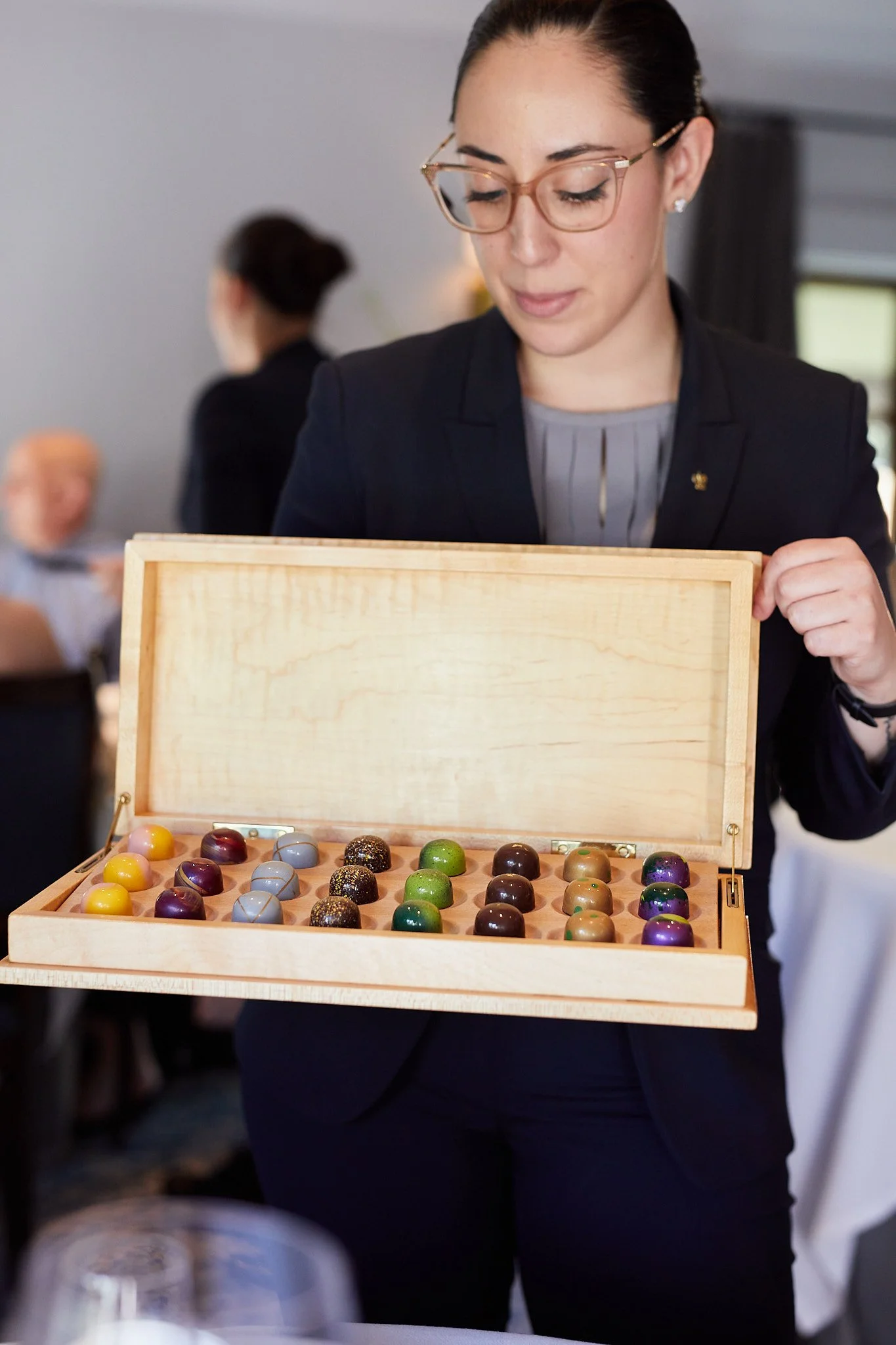 A woman with glasses and a black blazer is holding a wooden box filled with colorful, round chocolates at an event or gathering.