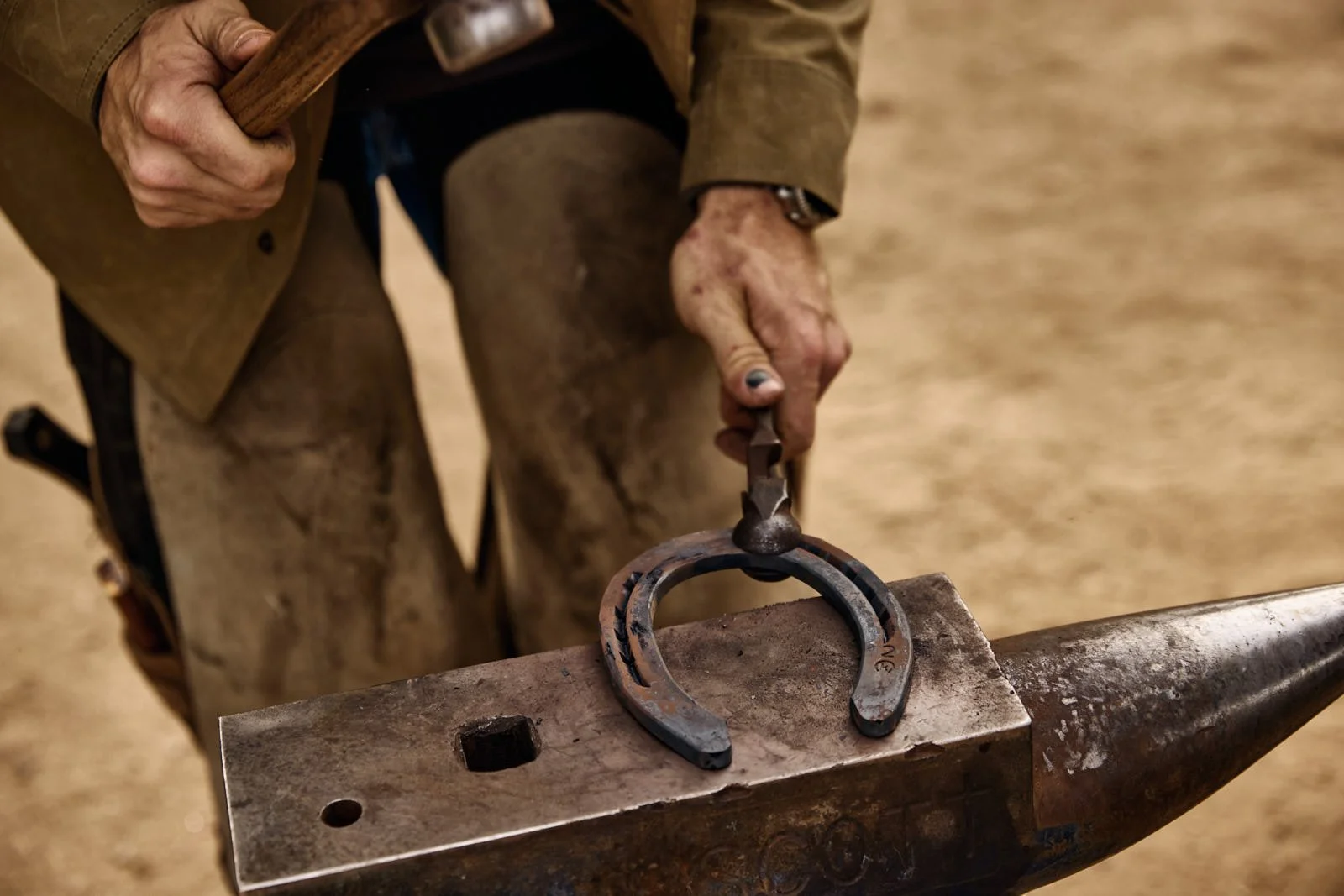 A blacksmith holding a horseshoe and using tools on a metal anvil.