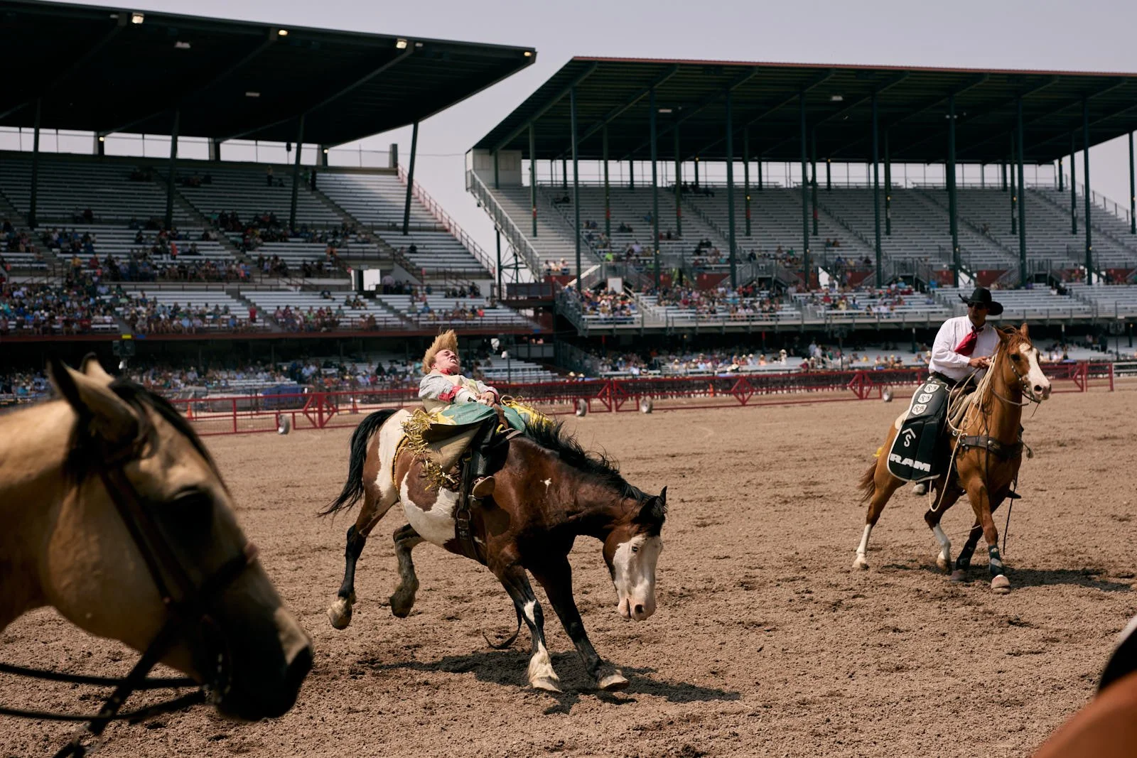 A rodeo scene with one rider on a bucking horse and another rider on horseback, with spectators in the grandstands in the background.