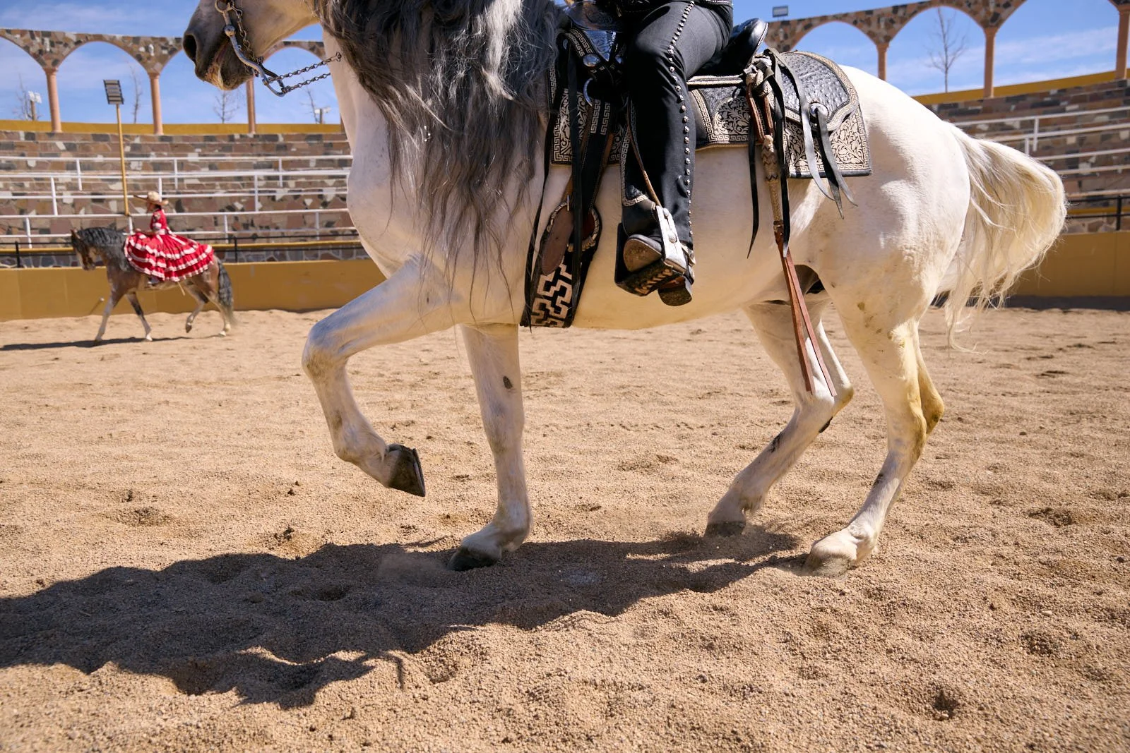 A white horse with a black mane and tail is being ridden in an arena with a light brown sandy surface. The horse has a saddle with intricate design and riding gear. In the background, a person in a red and white flamenco dress is riding another horse