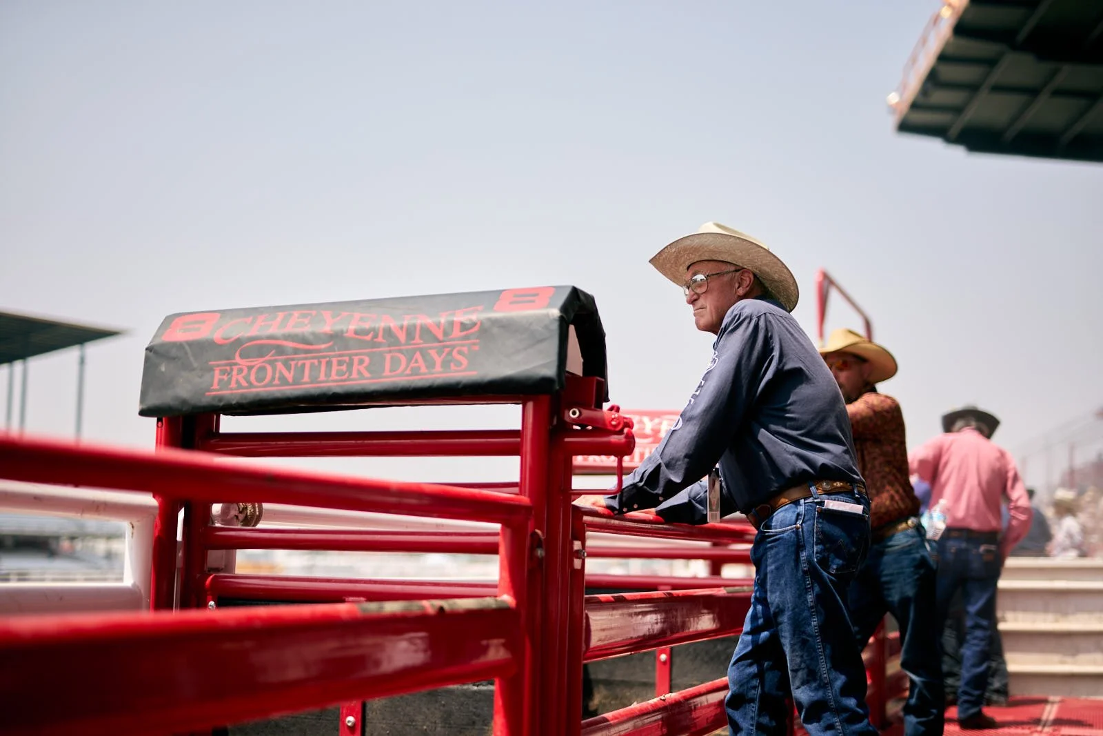 A man wearing a cowboy hat and glasses leans on a red railing at the Cheyenne Frontier Days rodeo event, with other men in cowboy hats in the background, under a clear sky.