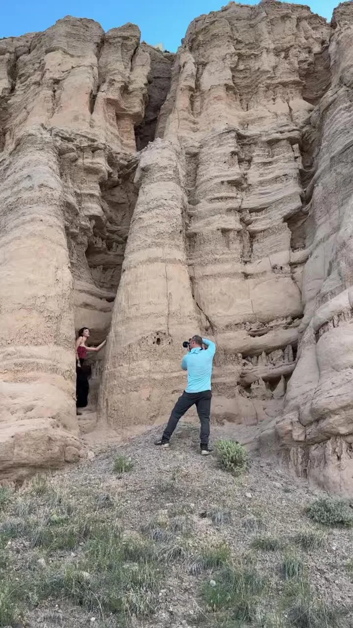 A woman standing next to large rock formations while John Robson photographs her in a desert landscape.