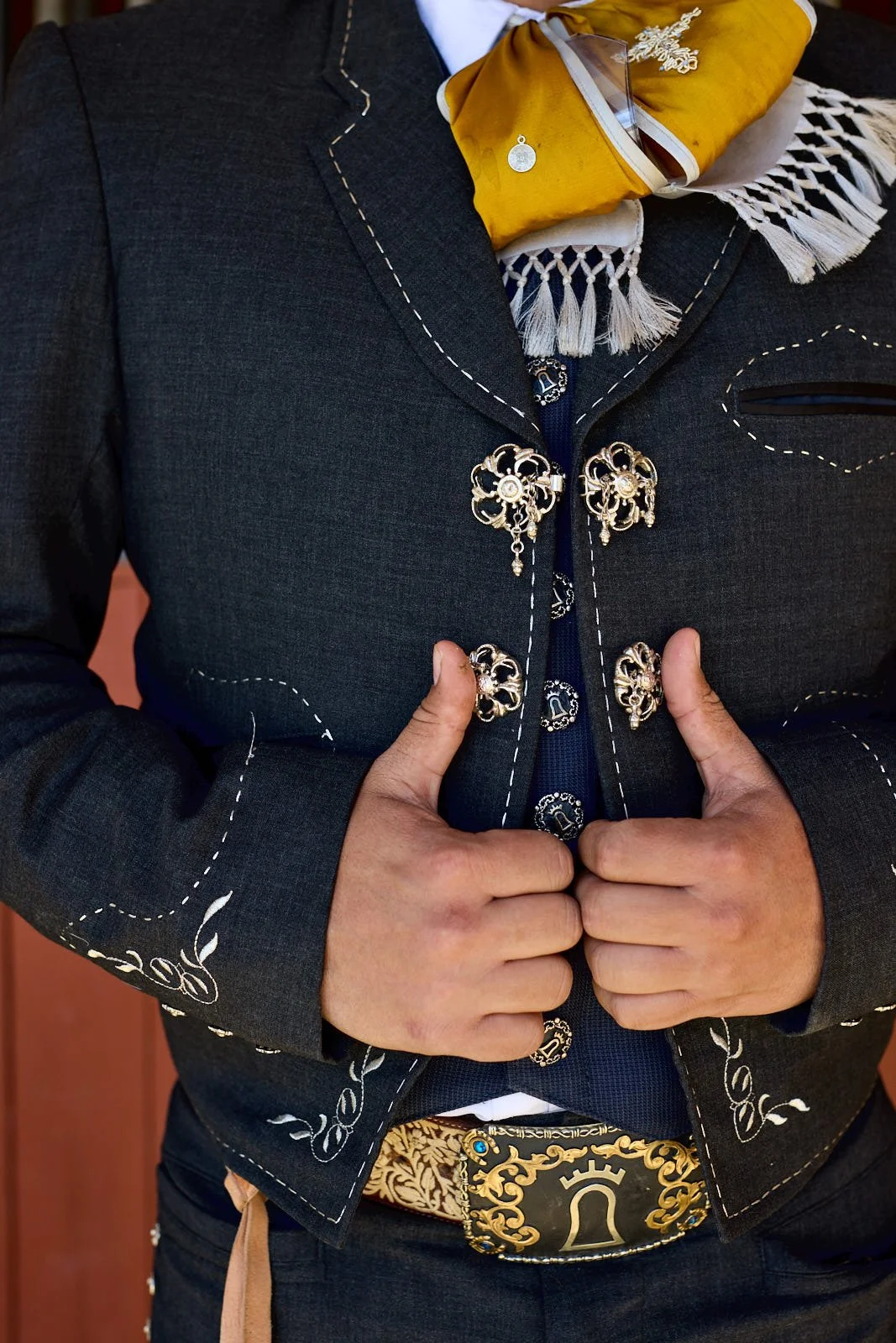 Close-up of a person wearing a dark Western-style jacket with decorative silver embellishments and stitching. The person is holding the front of the jacket with both hands, and their thumb is pointing inward, near a decorative belt buckle featuring a