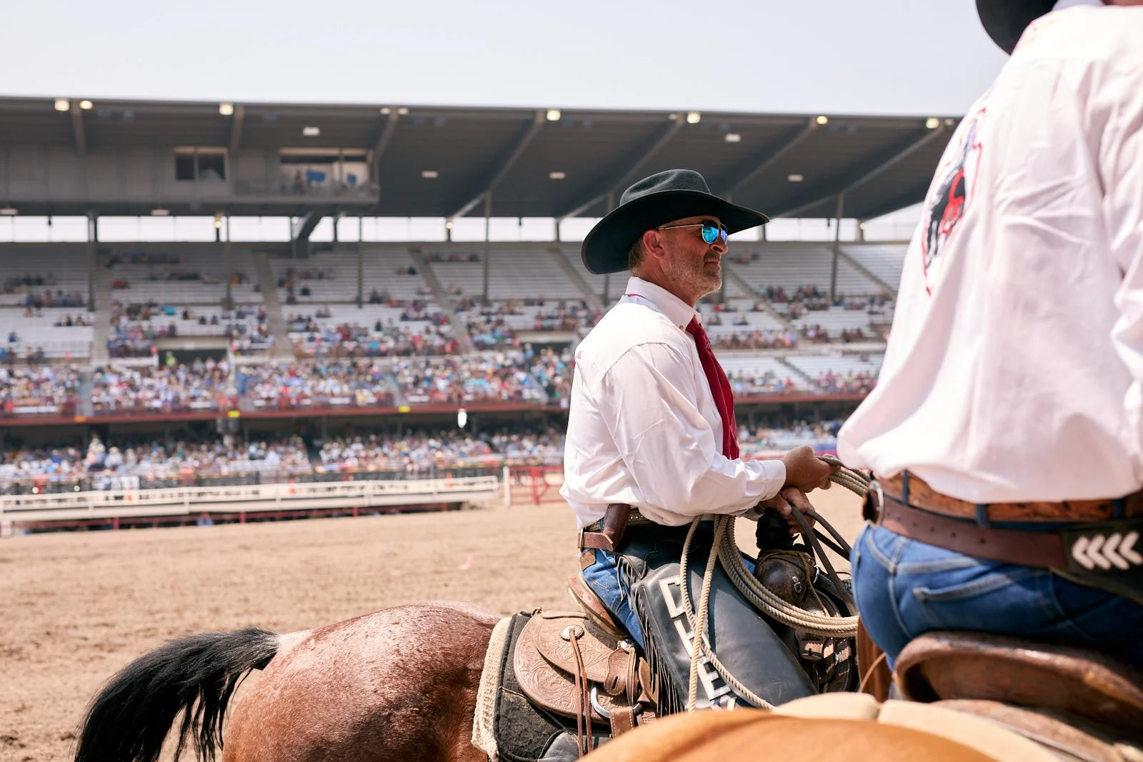 Two men in cowboy hats and white shirts on horseback in a rodeo arena, with a large crowd in the bleachers behind them.