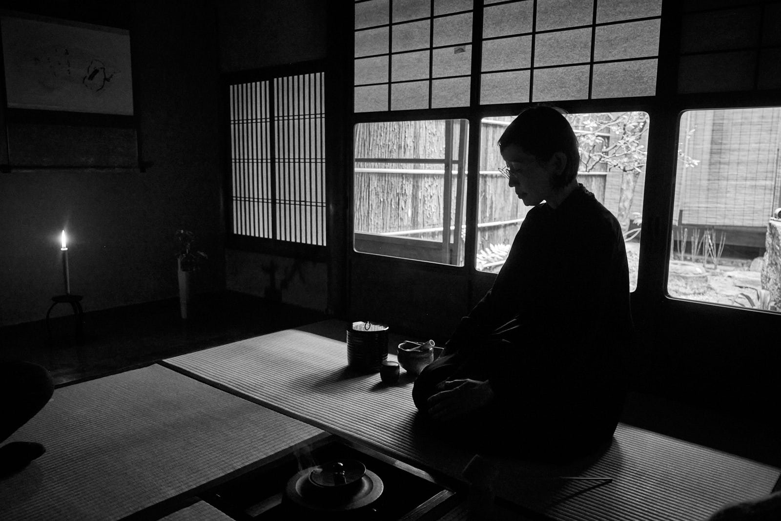 A person sitting in a traditional Japanese room with tatami mats, shoji screens, and a sliding door looking outside to a garden, with a lit candle on a small stand nearby.