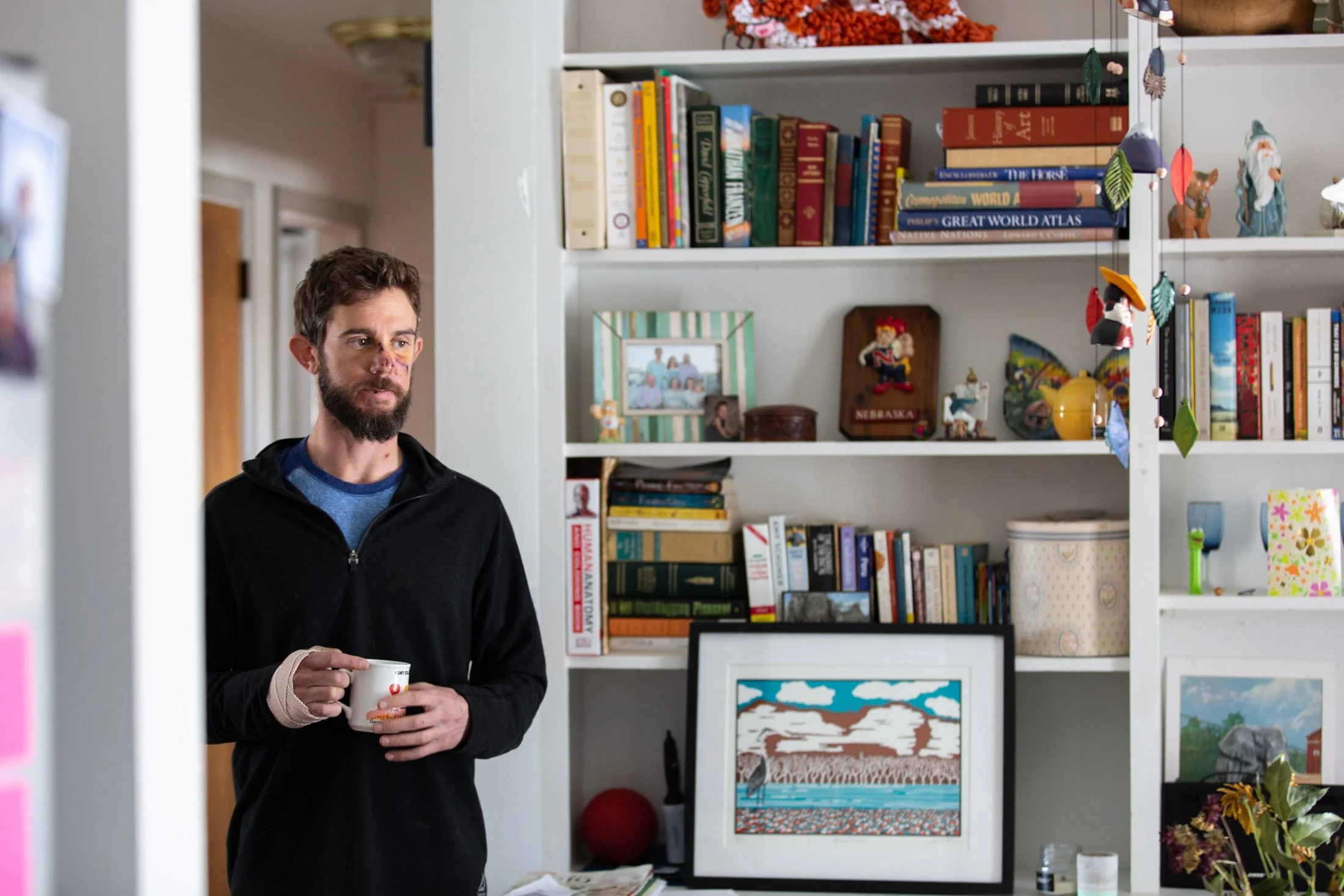 A man with a beard and casual clothing holding a mug in a room with a bookshelf filled with books and decorative items.