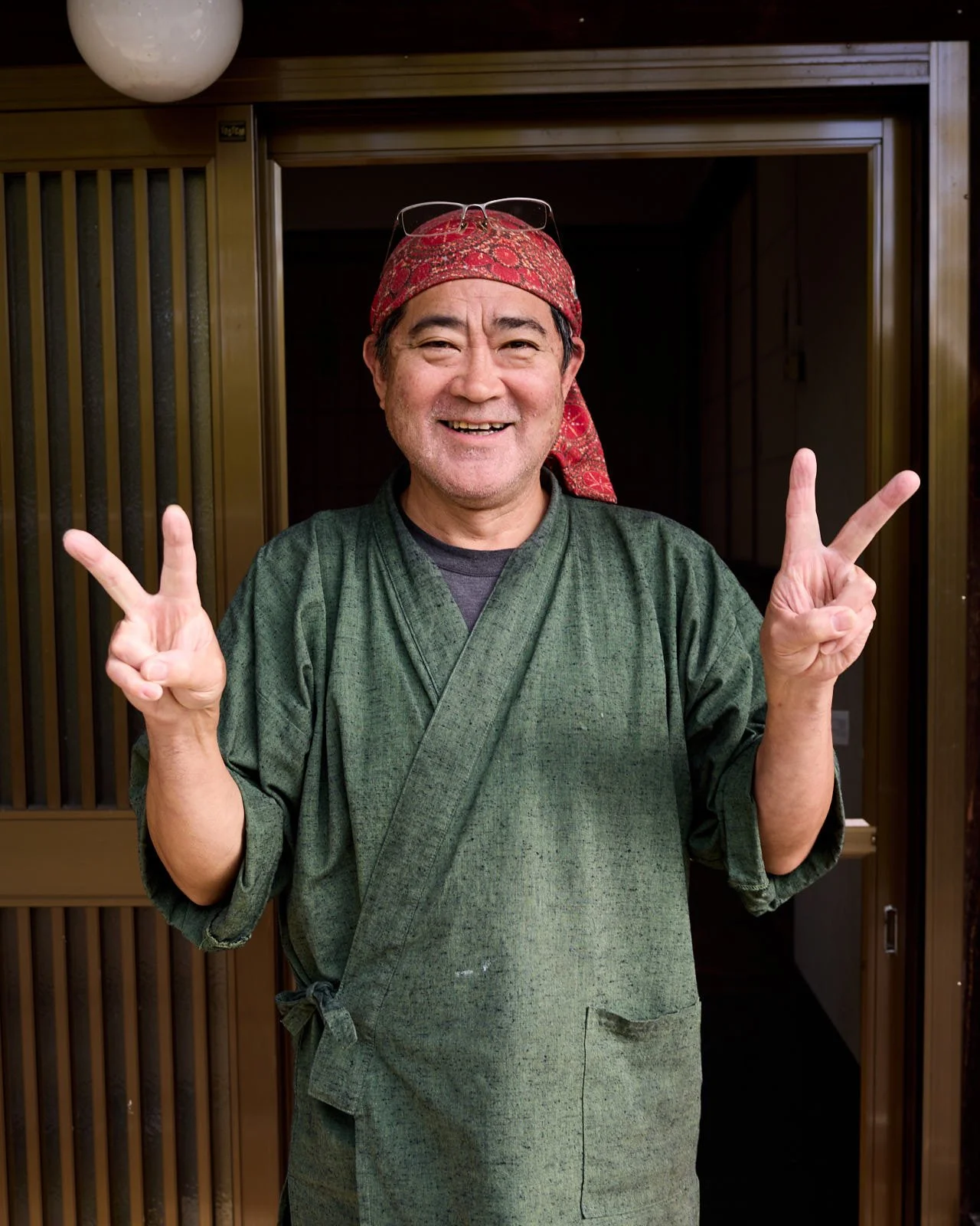A smiling man wearing a red bandana, glasses on top of his head, and a green traditional garment standing in front of a wooden door, making a peace sign with both hands.