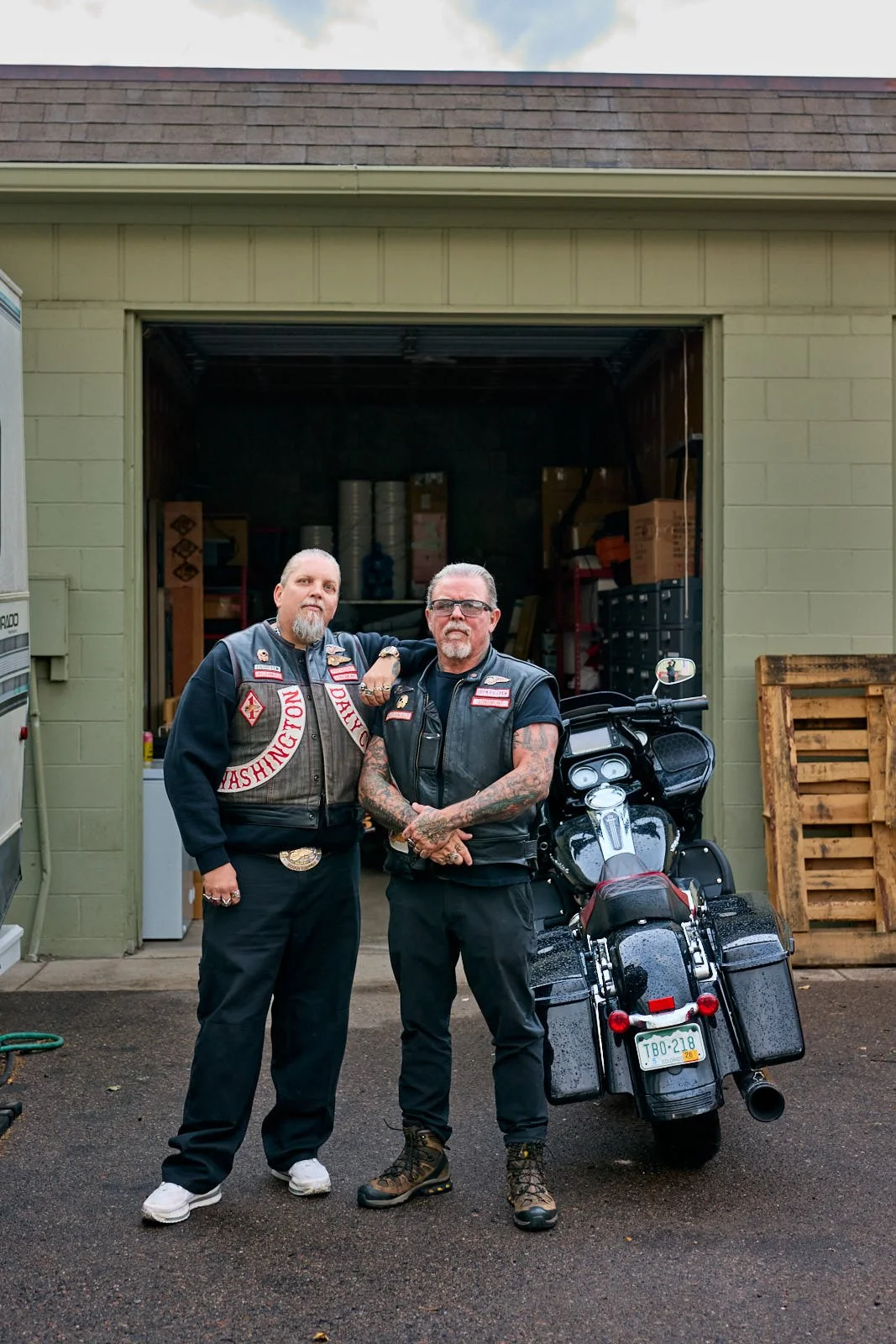 Two men standing in front of a garage with a motorcycle, both wearing black vests with patches, one with sunglasses and tattoos, the other with a beard, both posing for the photo.