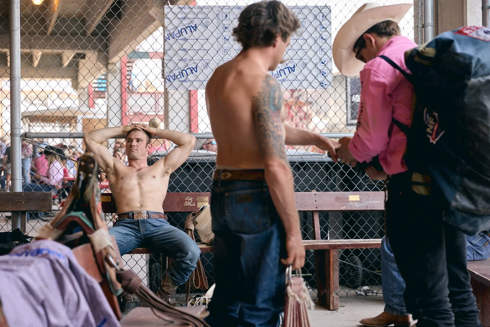 A young man with tattoos and no shirt sitting on a bench in a cowboy staging area at a rodeo