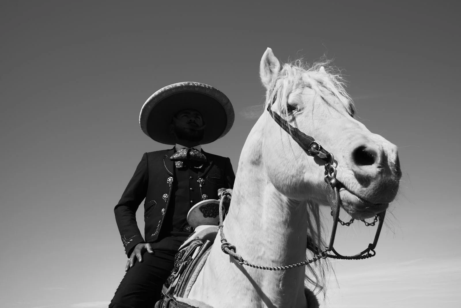 A man wearing a traditional Mexican outfit and sombrero, sitting on a white horse against a clear sky.
