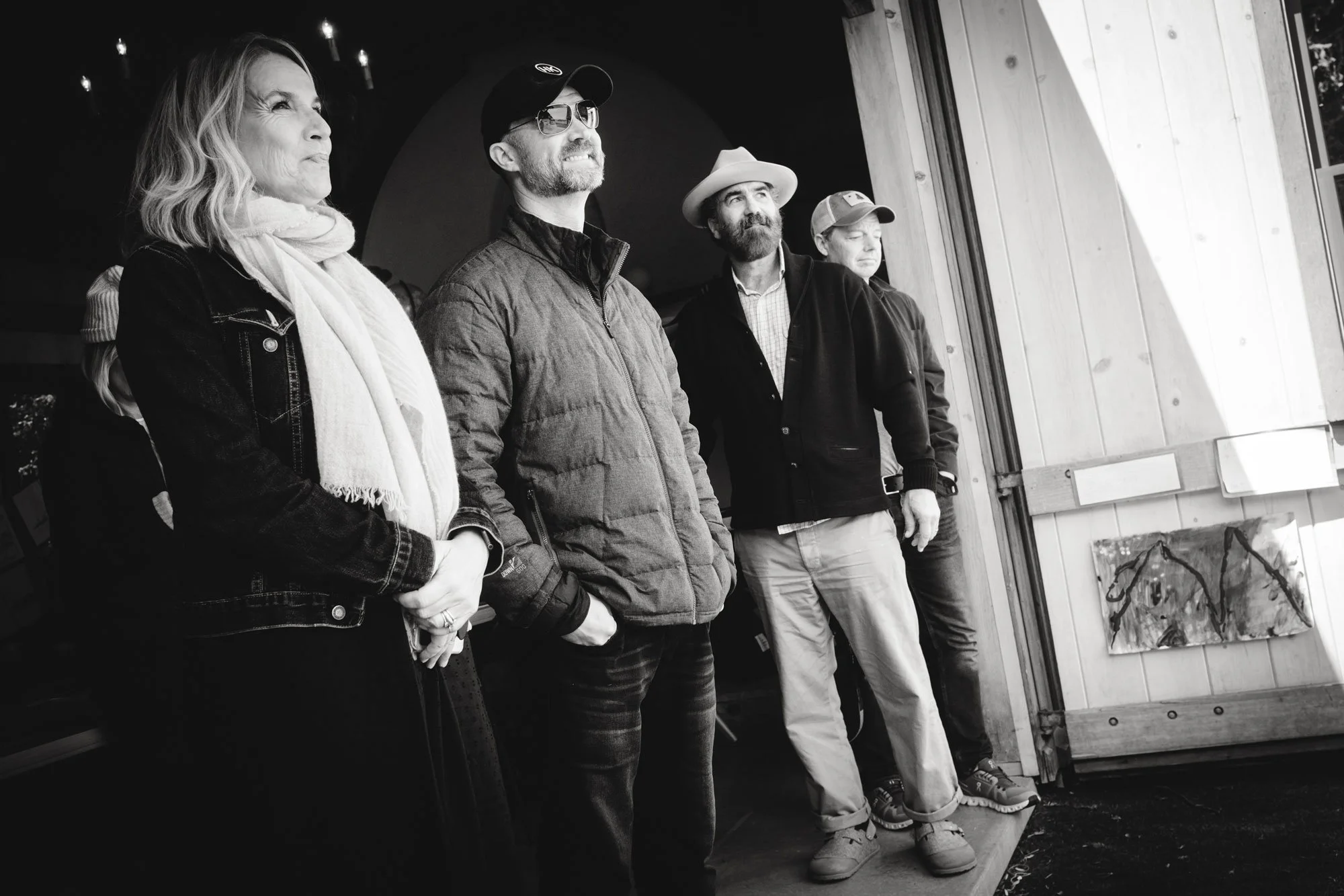 Four people standing in a row, looking to the right, in an indoor setting with wooden walls and art on display.