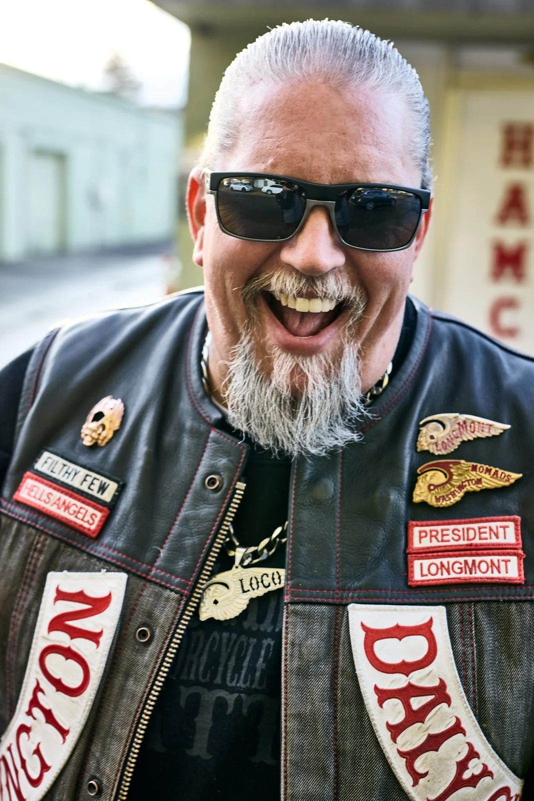 A happy man with sunglasses, a mustache, and a goatee wearing a leather vest with badges and patches, including one that says "PRESIDENT LONGMONT" and another that says "HELL'S ANGELS." He has a big smile, short gray hair, and is outdoors.