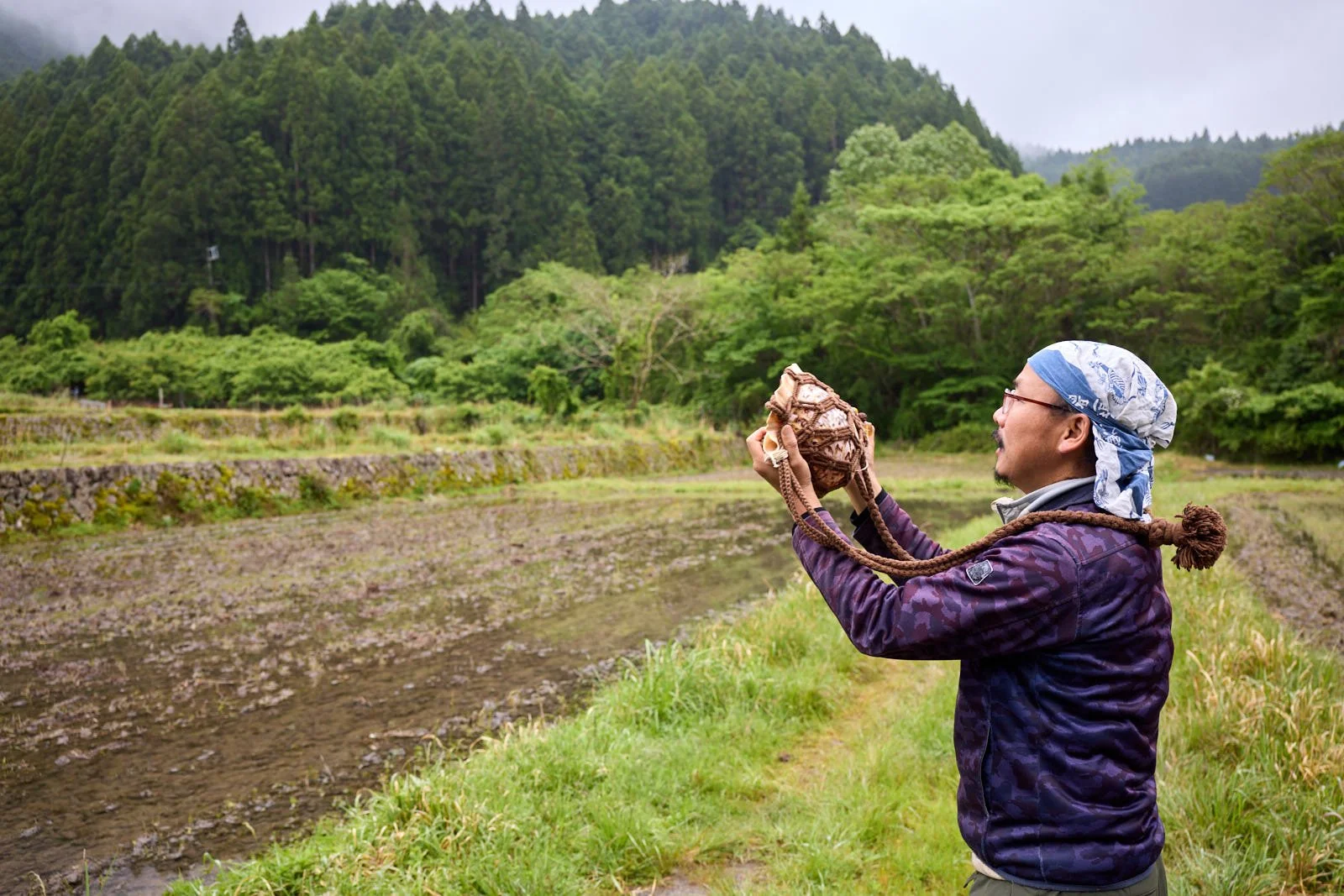 A man wearing a bandana, glasses, and outdoor clothing holds a conch shell with ropes in a rural landscape with green fields and forested hills.