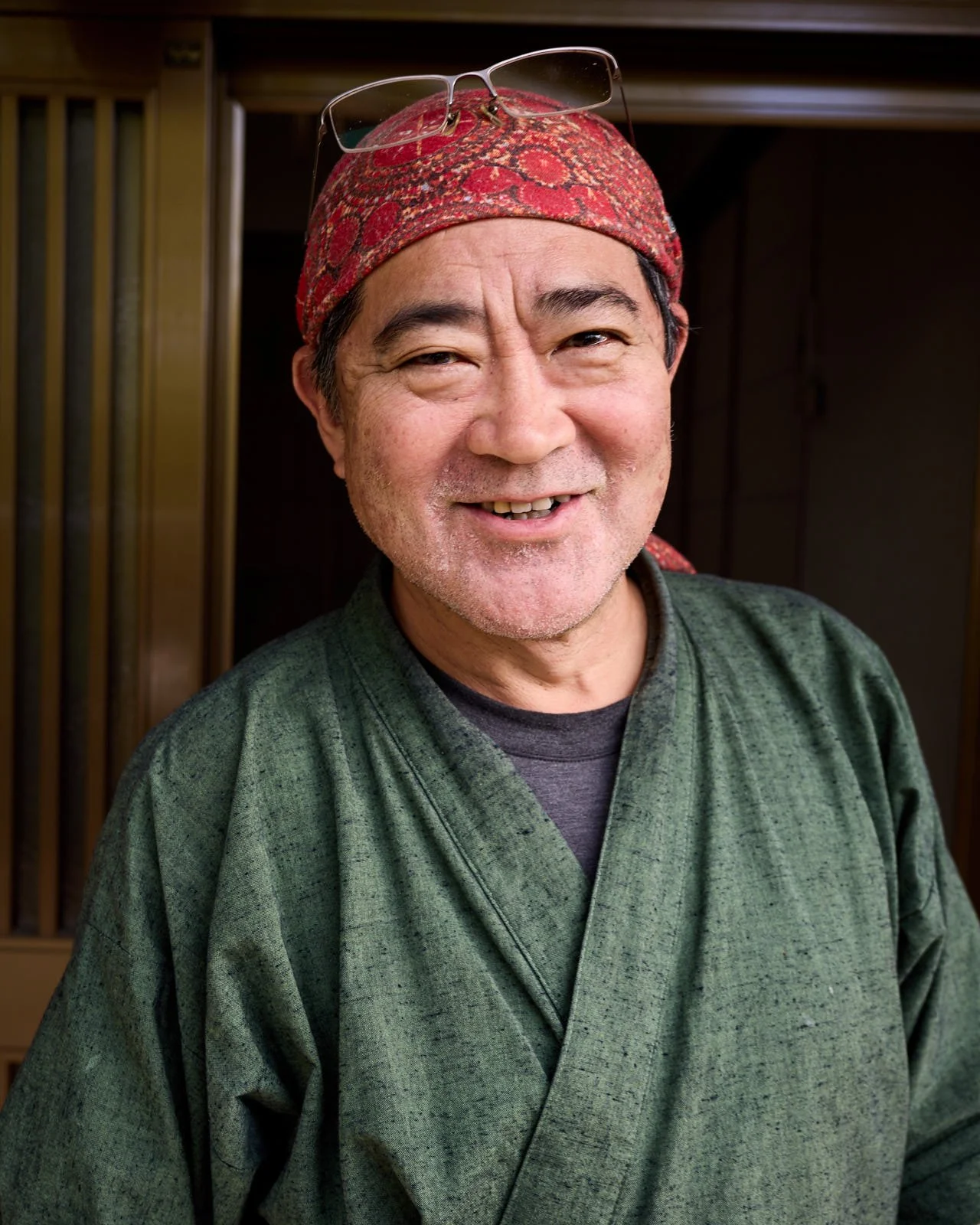 A smiling man wearing glasses on his head and a traditional Japanese kimono in front of a wooden door.