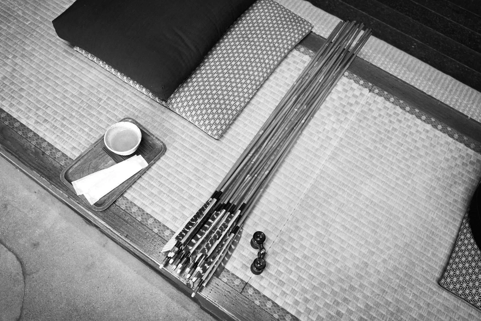 Traditional Japanese seating area with tatami mats, cushions, and a tray holding a cup and napkin, alongside a bundle of arrows and a pair of glasses.