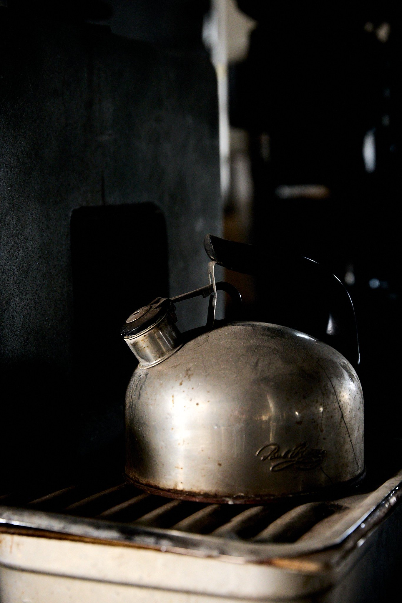 A vintage silver kettle resting on a stovetop in a dark workshop setting.