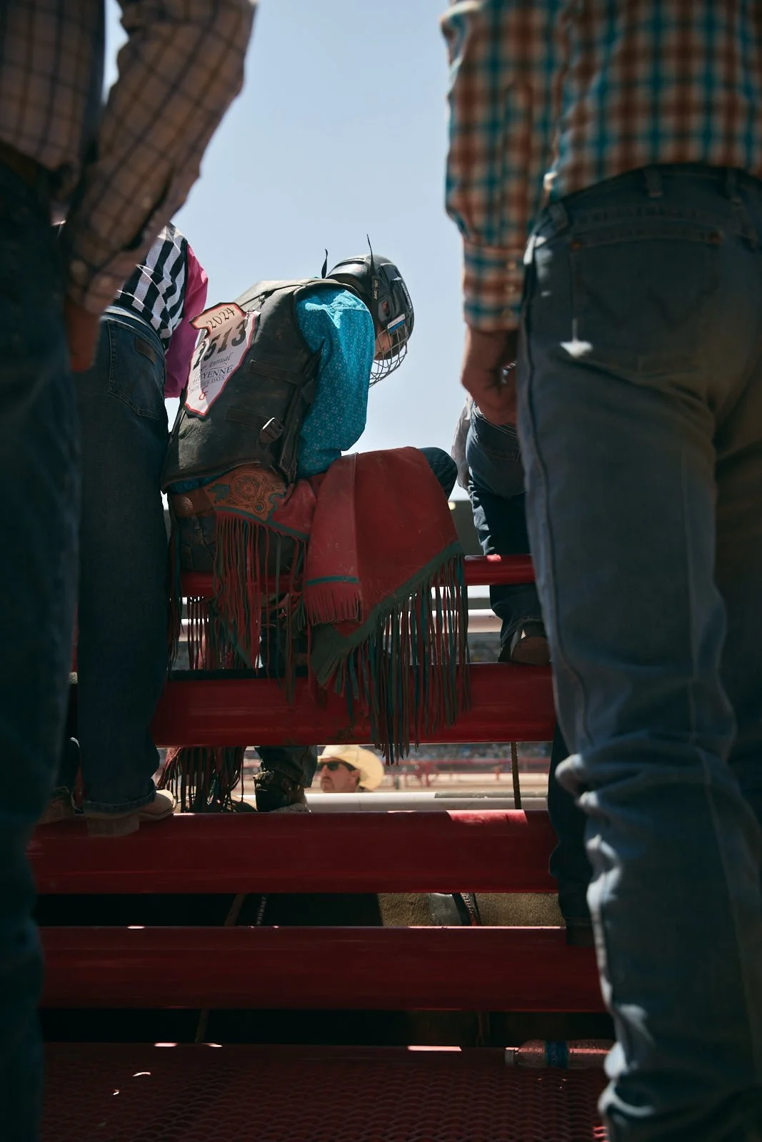 young bull rider in a blue shirt, black helmet, sitting on chute rail at a rodeo