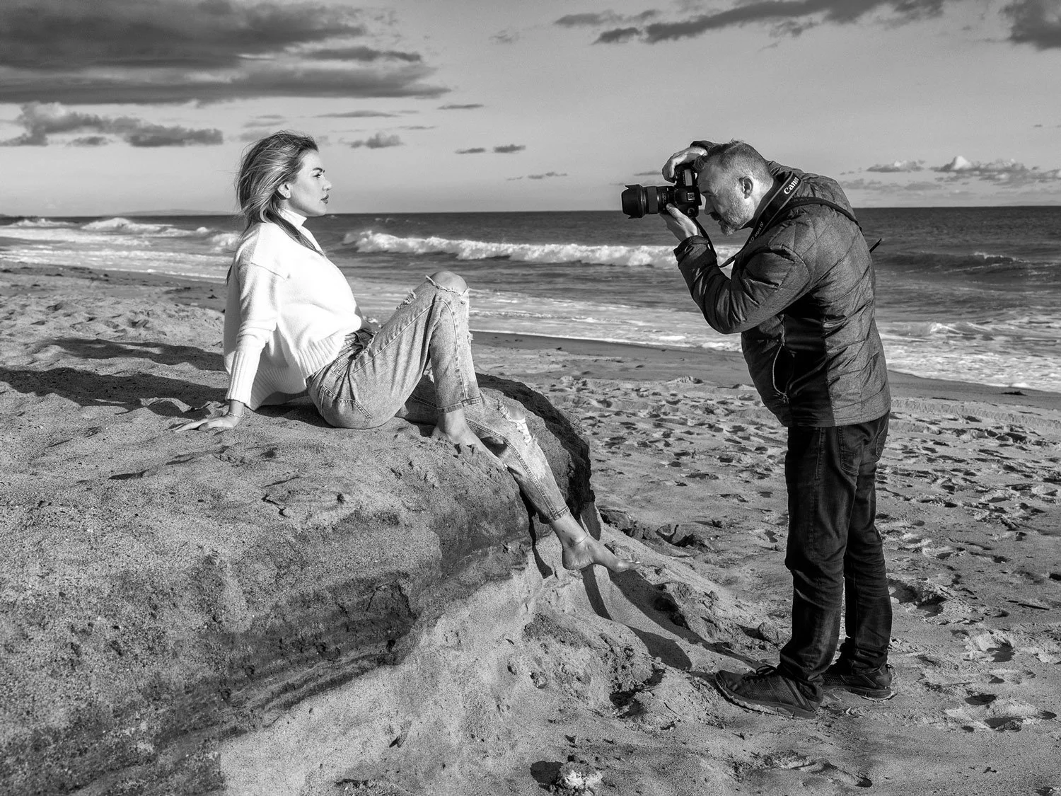 A woman sitting on a large rock a California beach, posing for photographer John Robson taking her picture. The scene is outdoors under a partly cloudy sky with ocean waves in the background.