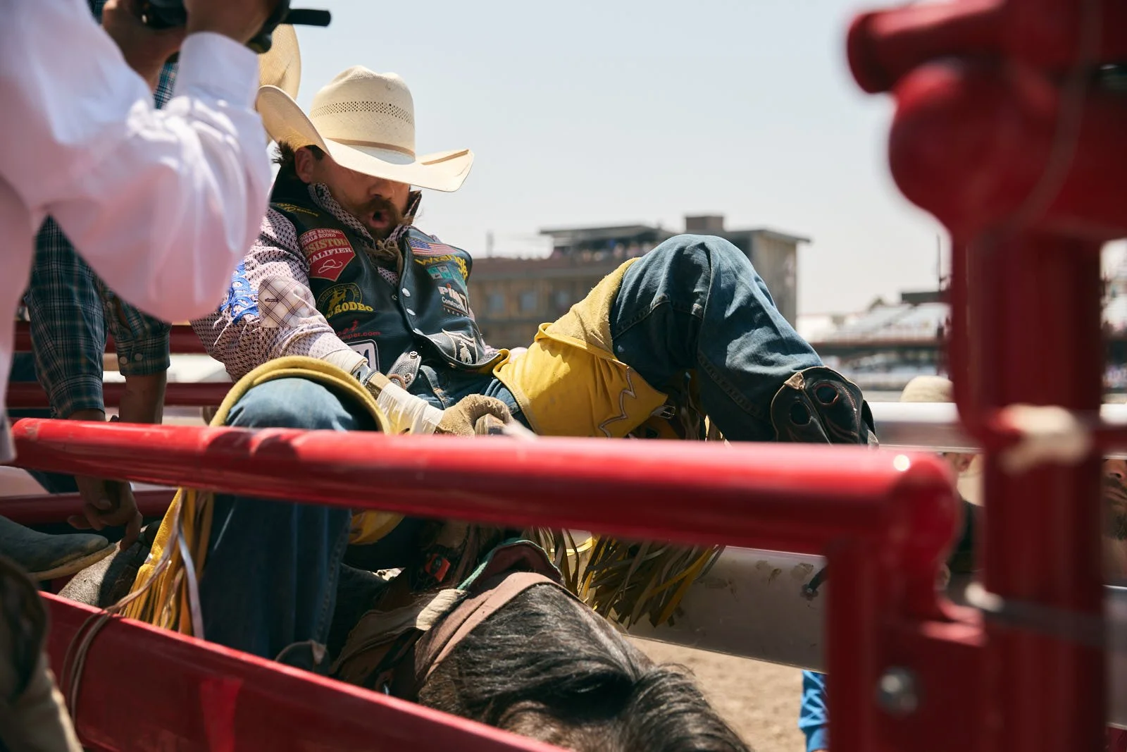 A rodeo cowboy on a bull, surrounded by other people, one wearing a cowboy hat. They are all near a red metal chute on a sunny day with areana in the background.