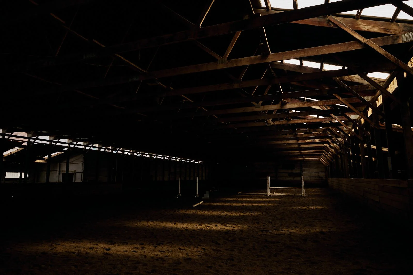 Interior view of an empty indoor horse riding arena with a dirt floor and wooden support beams, partially illuminated by natural light.