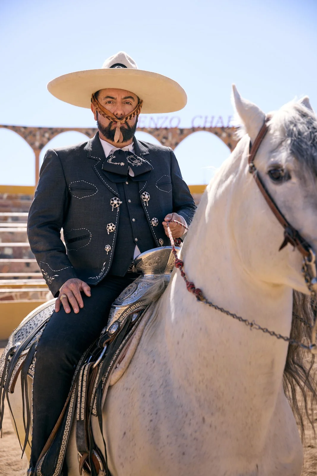 A man dressed in traditional Mexican charro attire, riding a white horse at an outdoor arena under a clear blue sky.