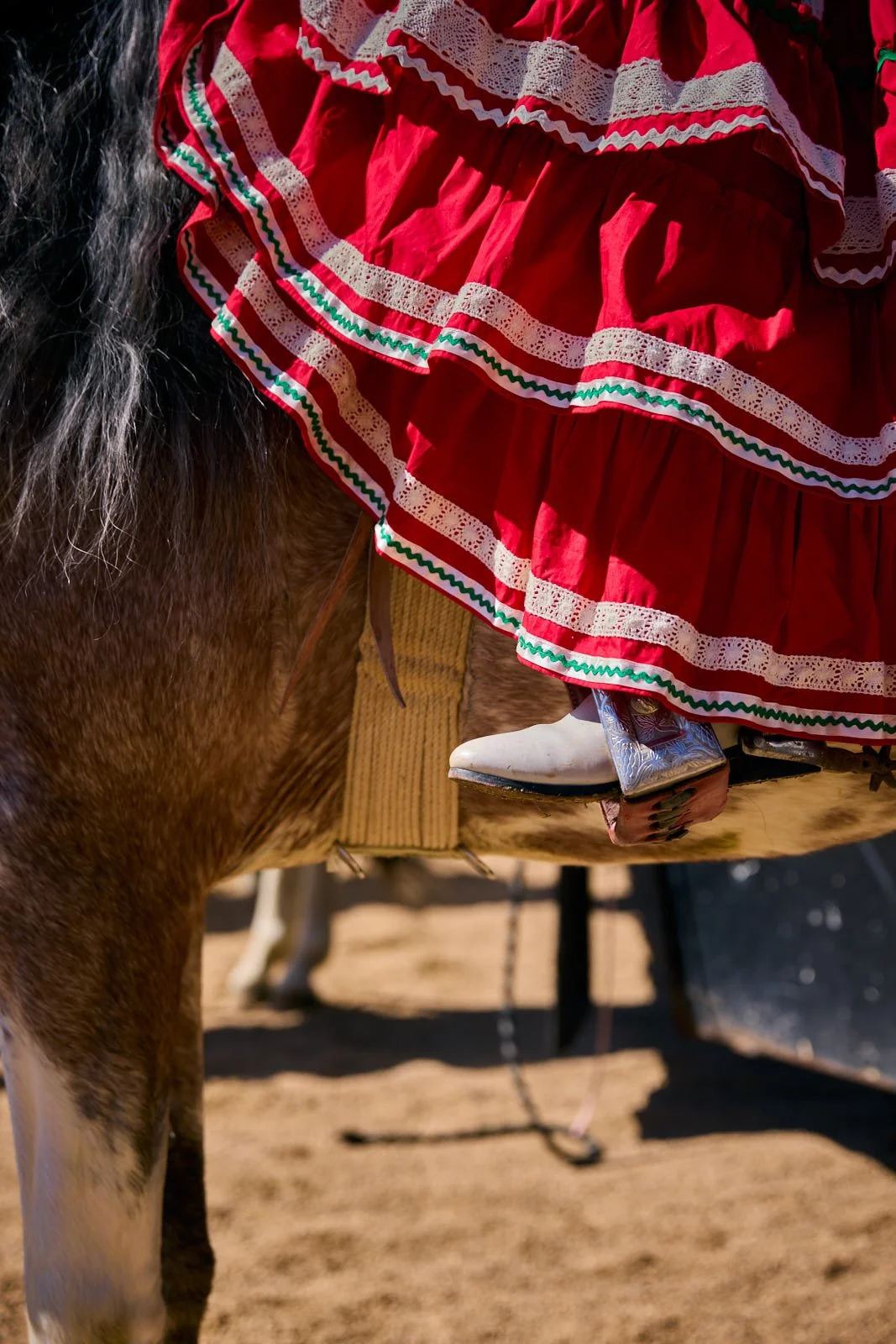 Close-up of a person dressed in a traditional red dress with white lace and green accents, sitting on a brown horse with a beige saddle, with sandy ground in the background.