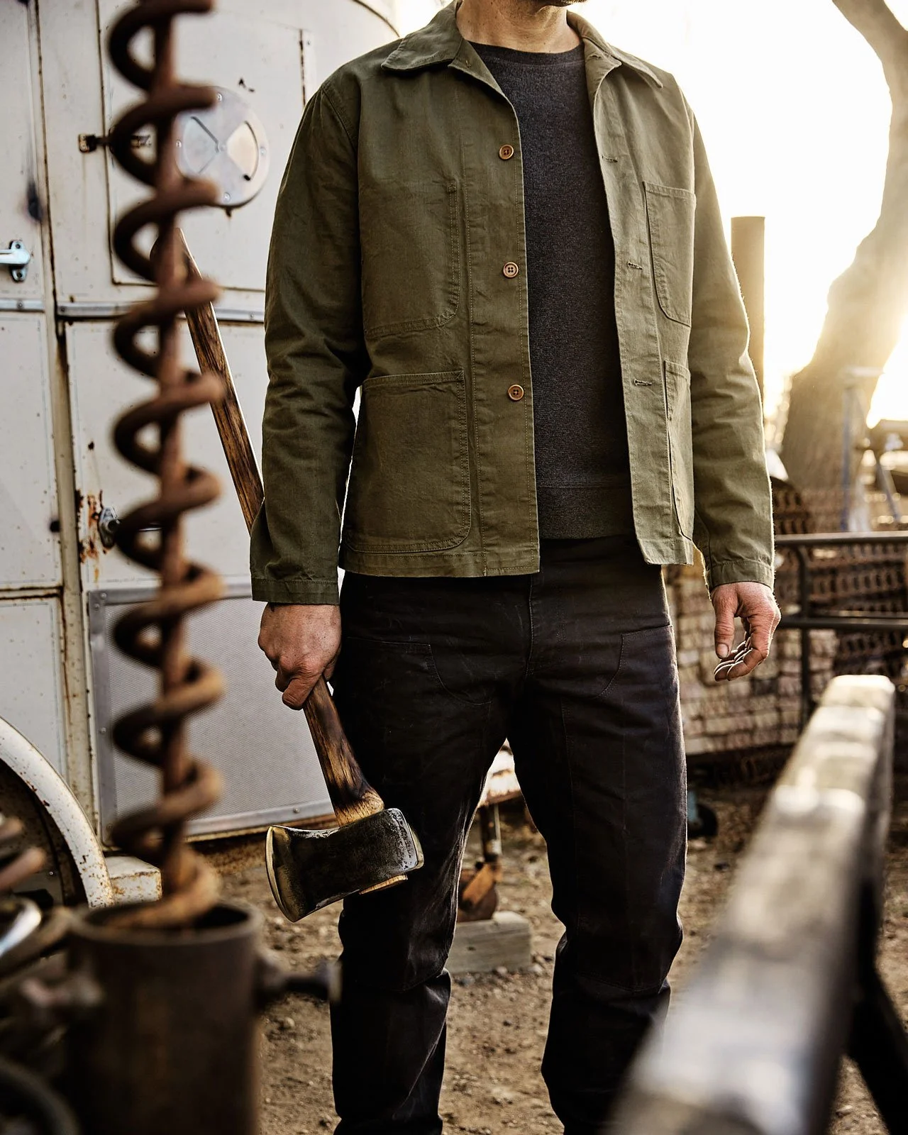 A person standing outdoors holding an axe, wearing a green jacket and black pants with sunlight shining through in the distance.