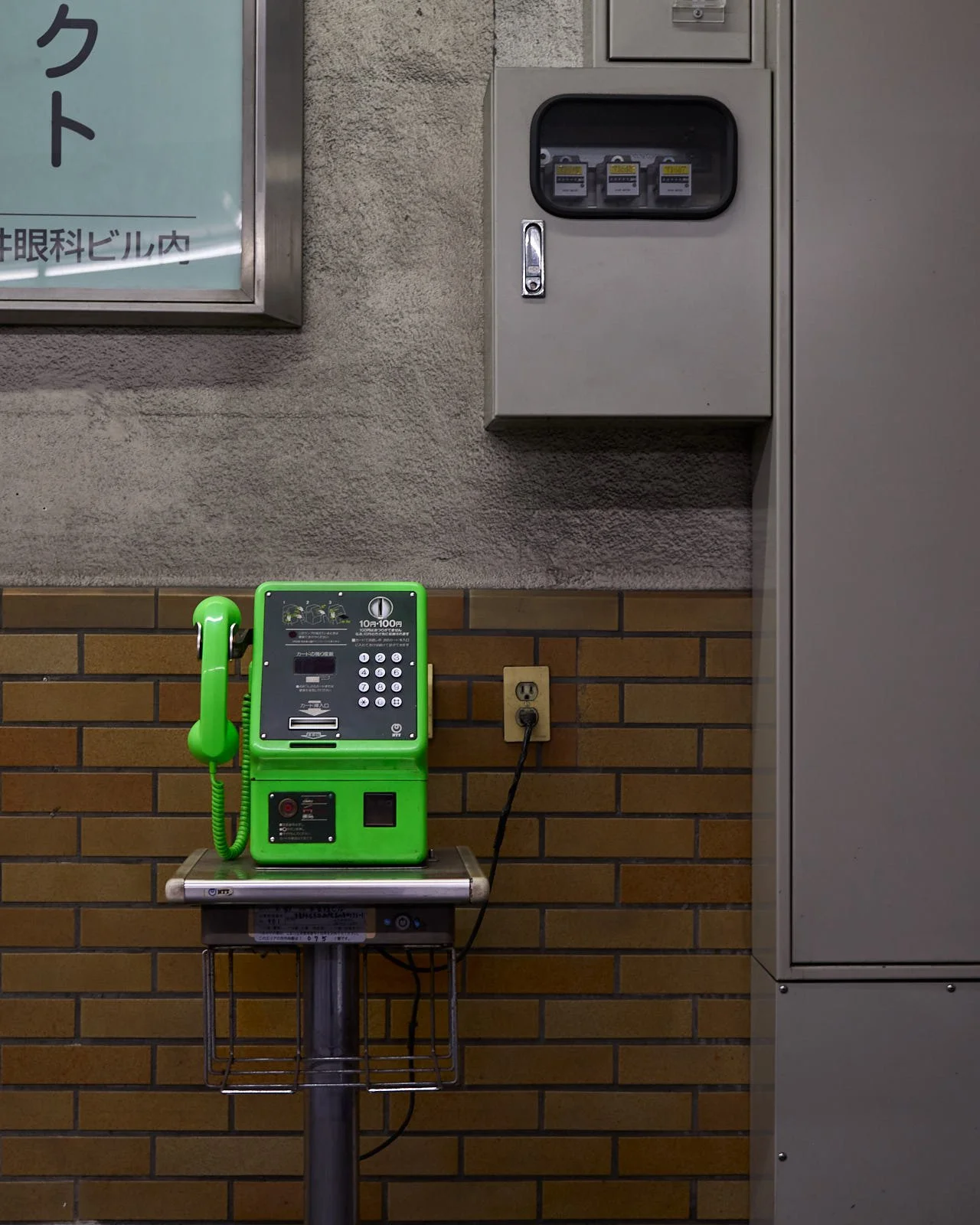 A bright green public telephone with a coin slot and keypad, placed on a metal stand against a brick wall.