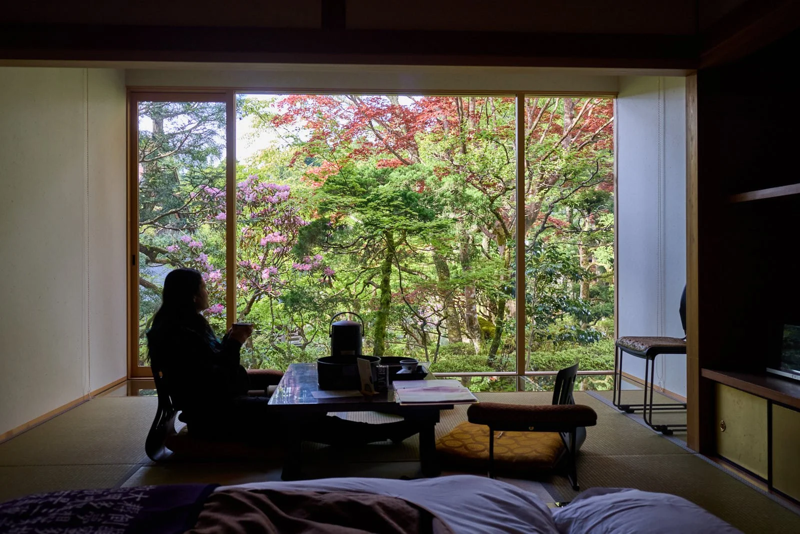 A woman sitting on a cushion and using a smartphone near a low table in a traditional Japanese room with a large window overlooking a lush garden with blooming trees.