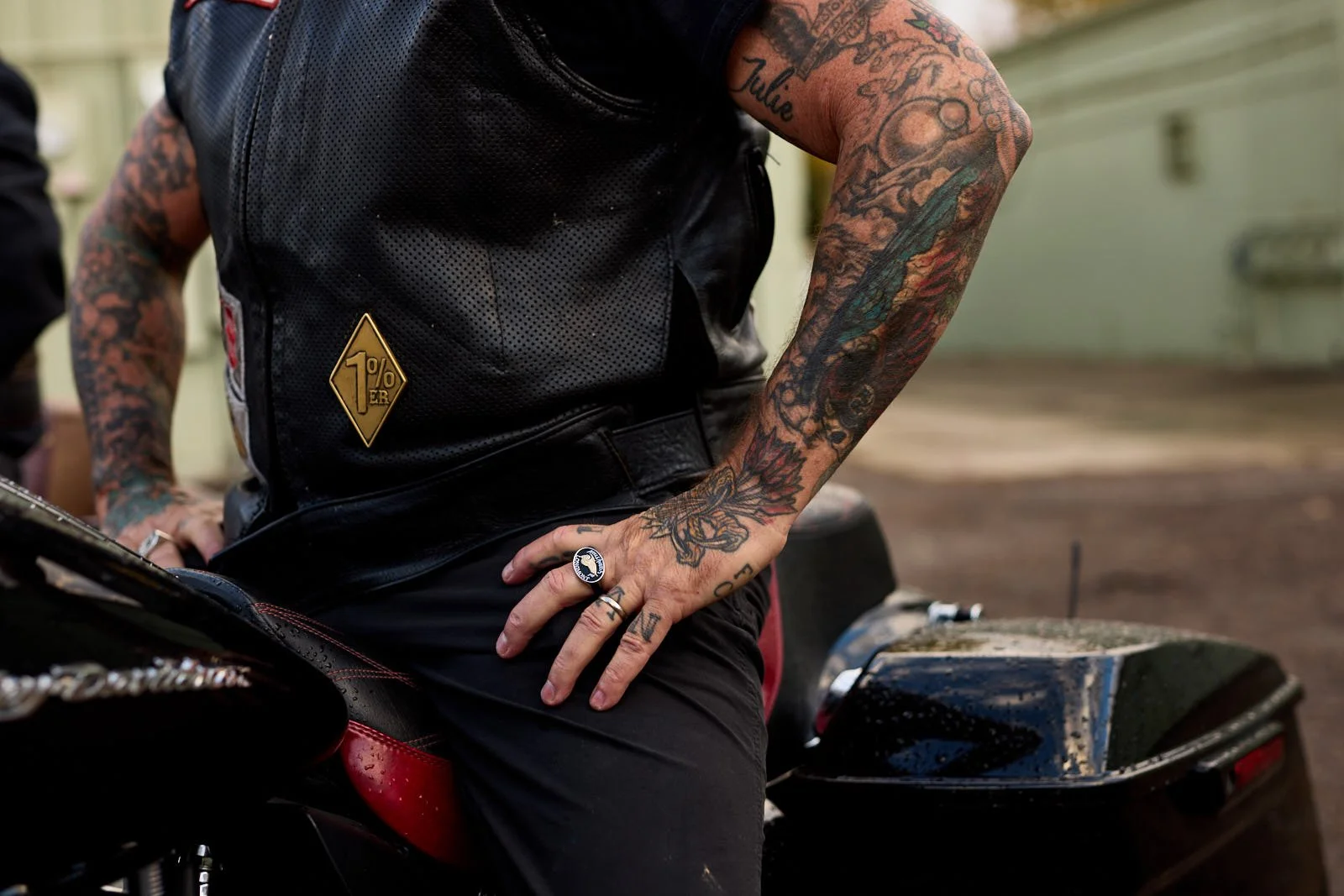 Person with tattoos wearing a black leather vest and rings, sitting on a motorcycle.