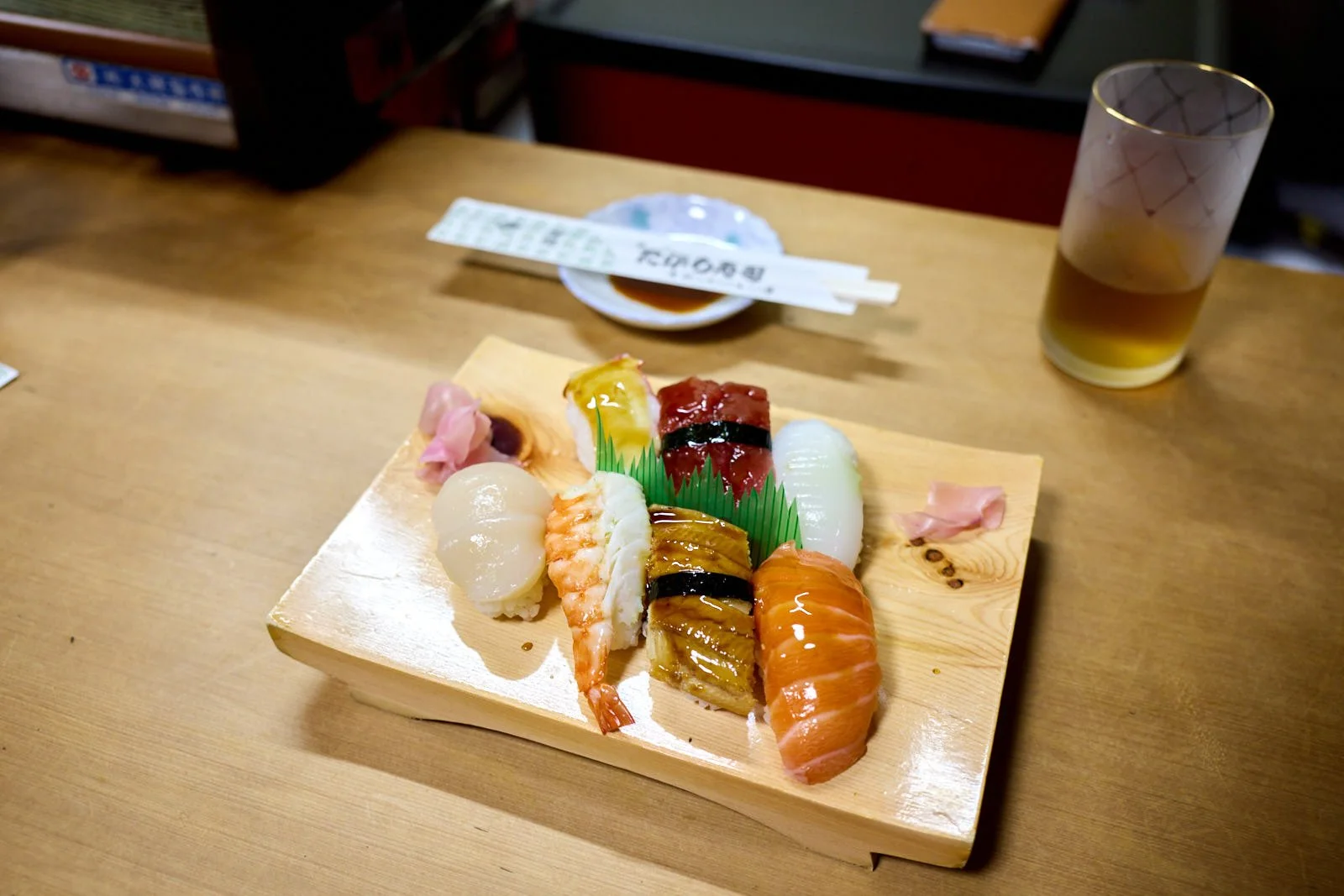 Sushi platter with various types of sushi including sashimi, nigiri, and rolls, served on a wooden platter on a restaurant table with a glass of beer and soy sauce.
