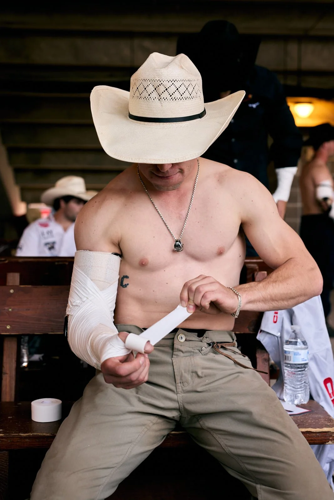 A shirtless man with a cowboy hat sitting on a wooden bench, wrapping his arm with a bandage.