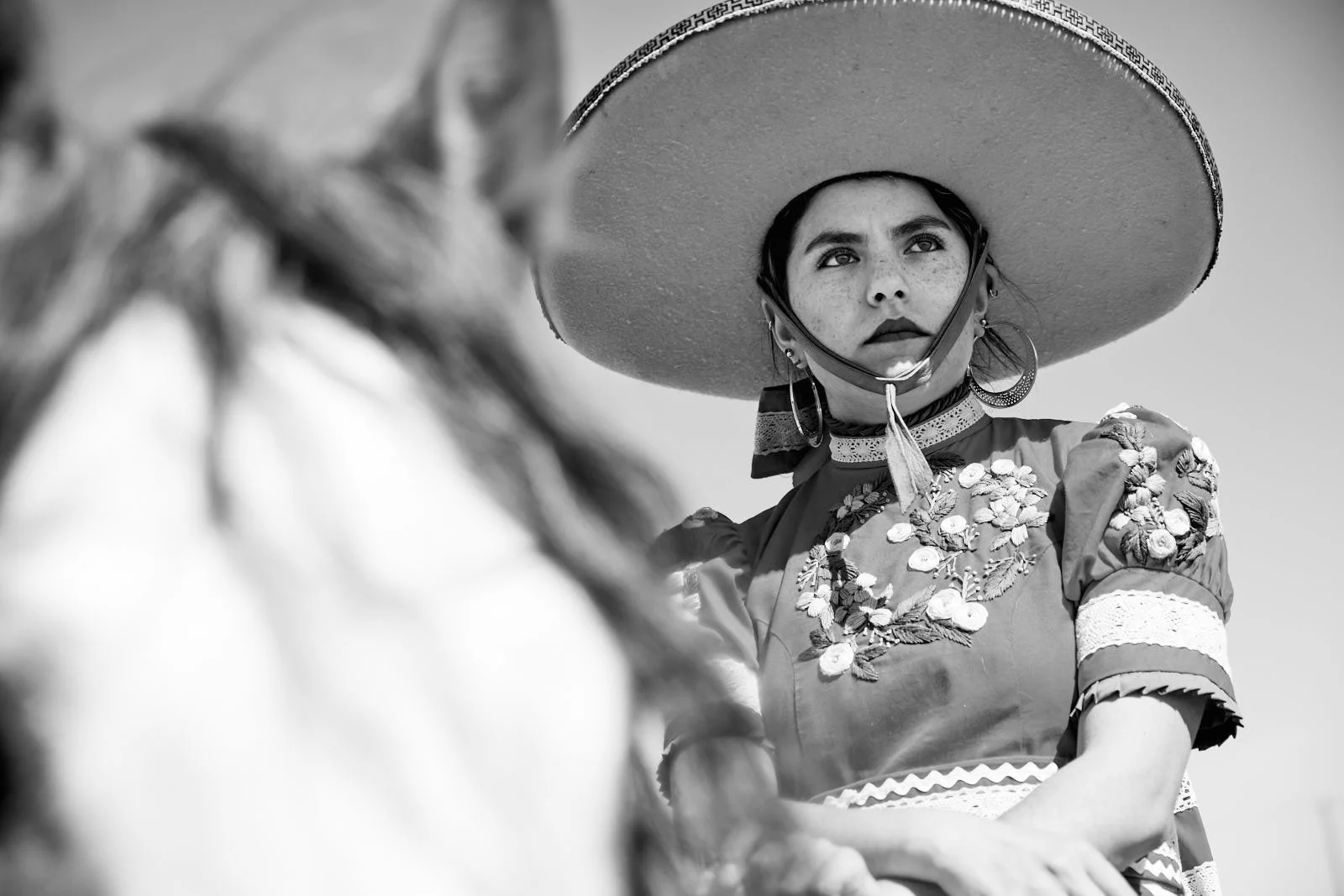 A young woman wearing a large sombrero, hoop earrings, and a floral embroidered dress looks into the distance with a serious expression.